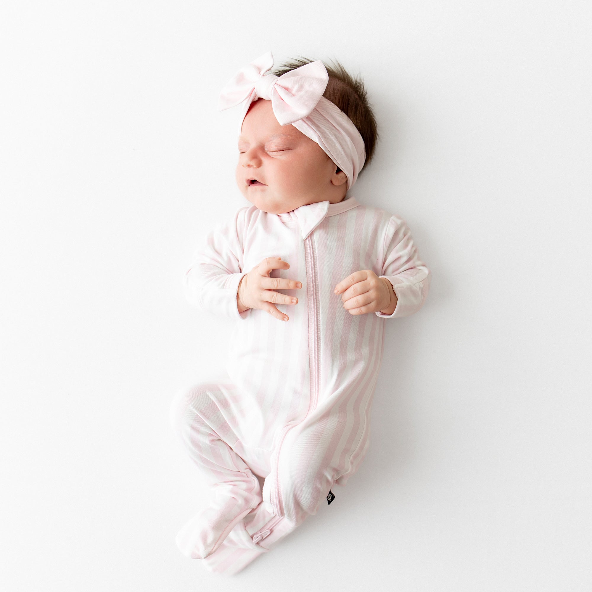 Sleeping newborn wearing the Zippered Footie in Small Sakura Stripe and Light pink bow headband