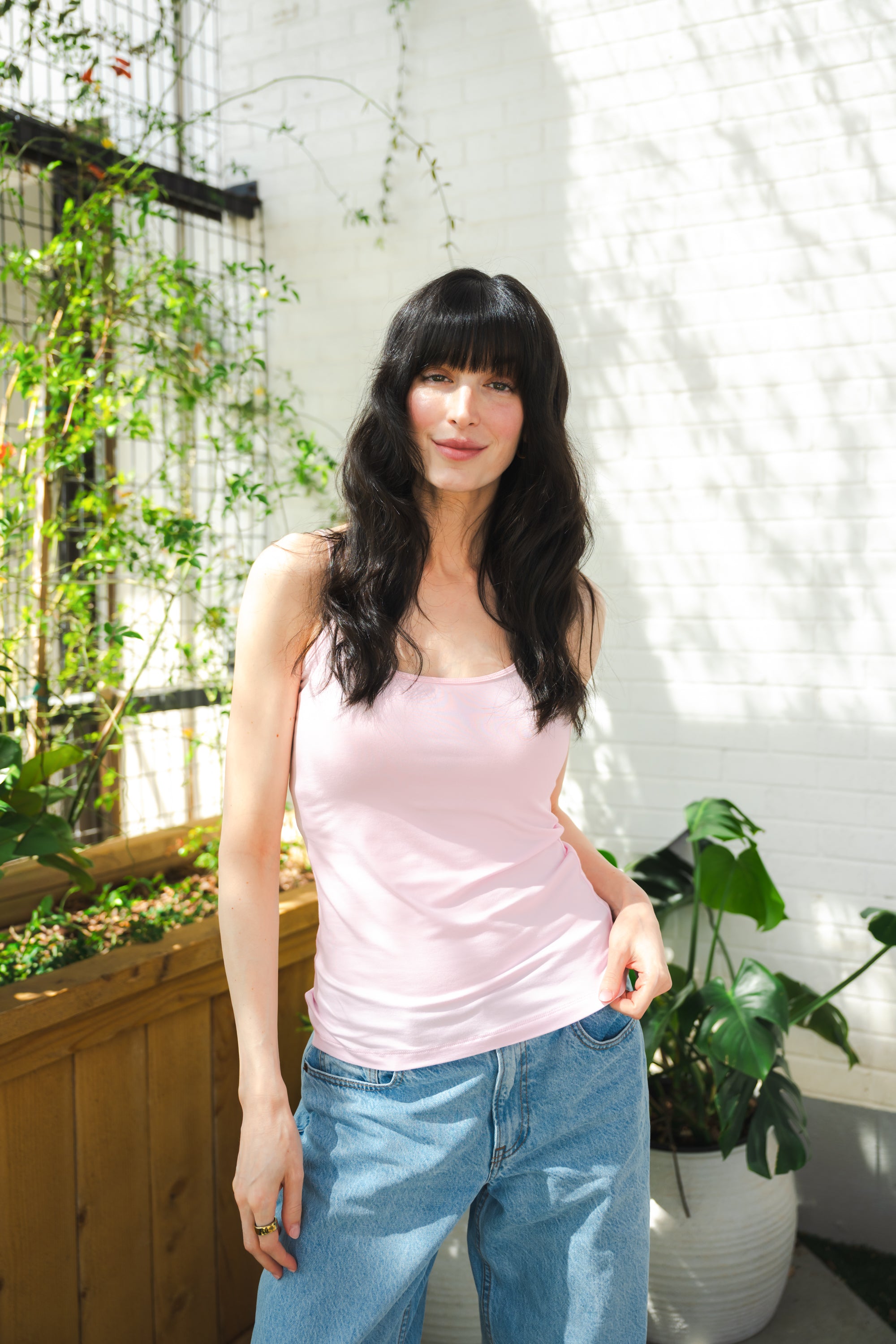Woman wearing a pink tank top and blue jeans standing in a bright indoor setting with plants.