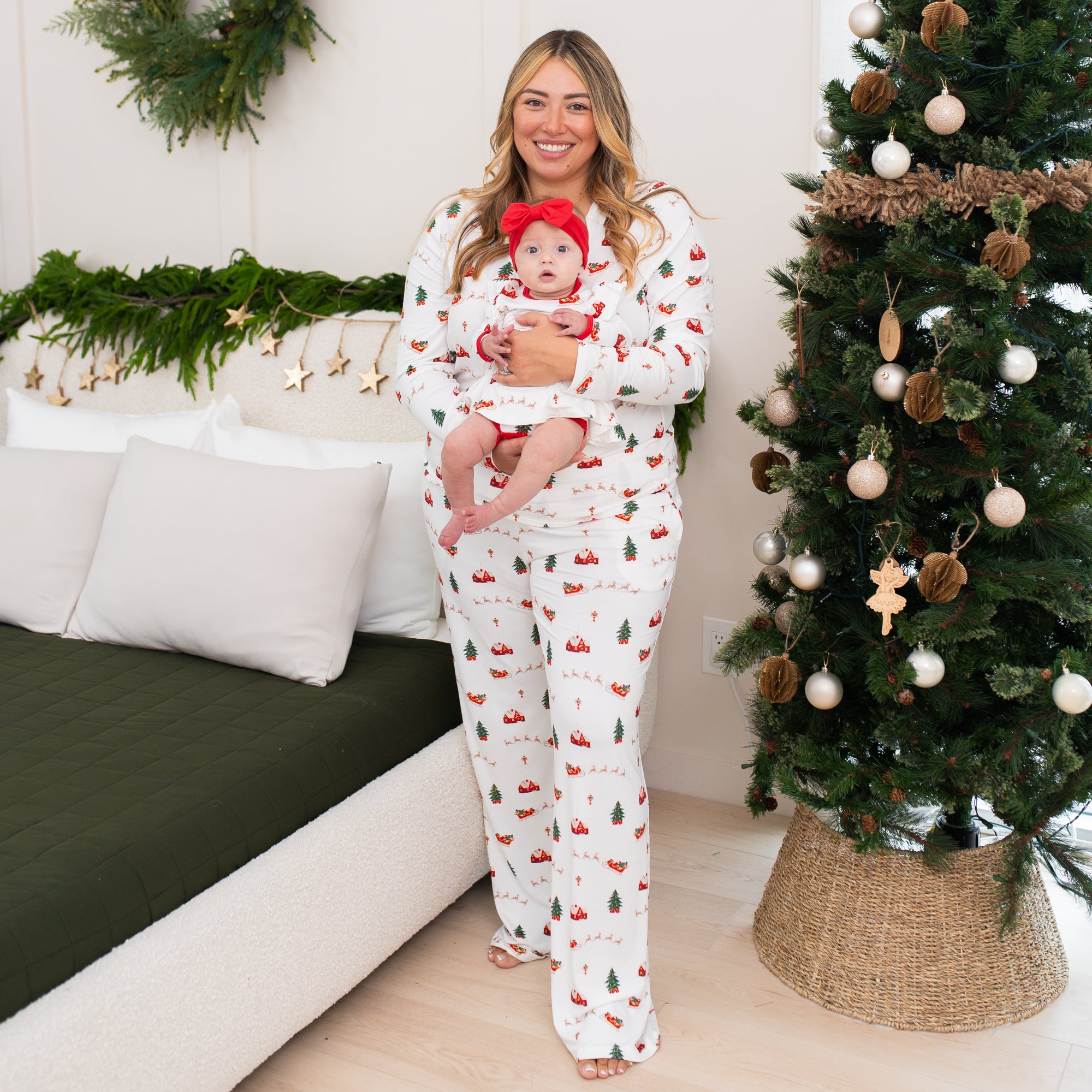 Mother standing between a bad and christmas tree holding her infant daughter both matching in Santa Sleigh