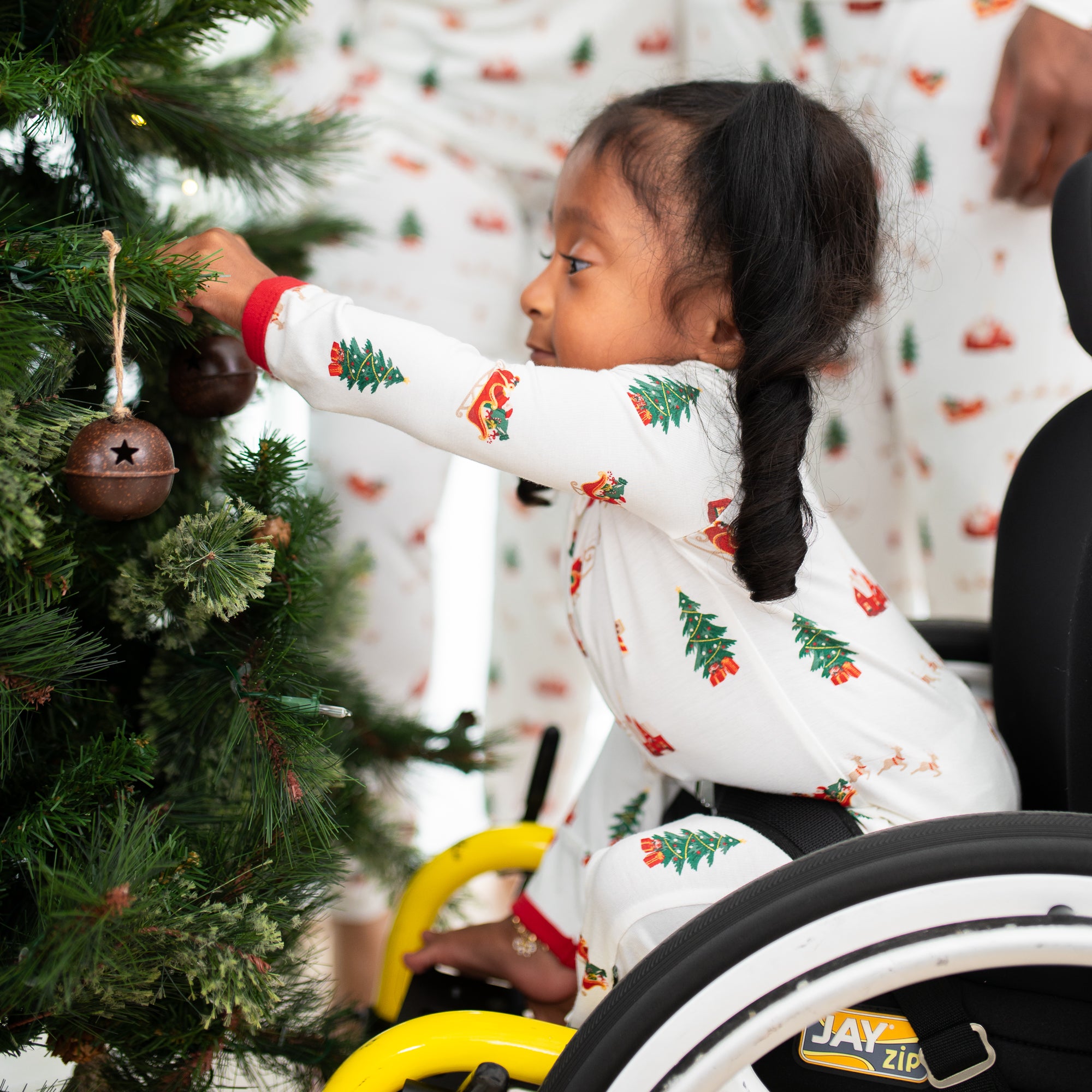Young girl wearing the Zippered Romper in Santa Sleigh putting an ornament on a christmas tree