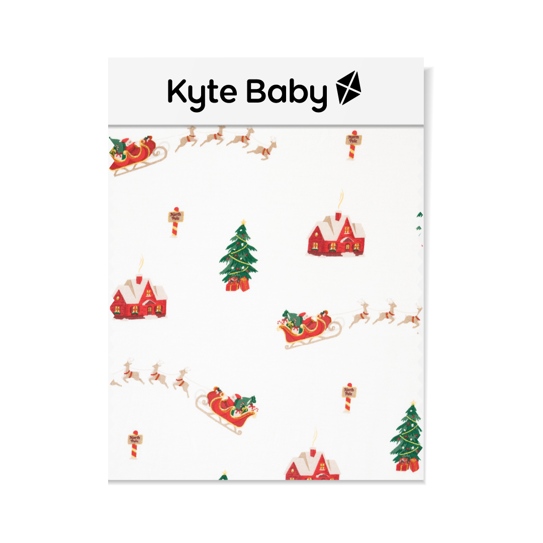 A swatch of Kyte Baby's Santa Sleigh print featuring a Christmas Tree, north pole sign, a santa sleigh with reindeers and red house on a white background