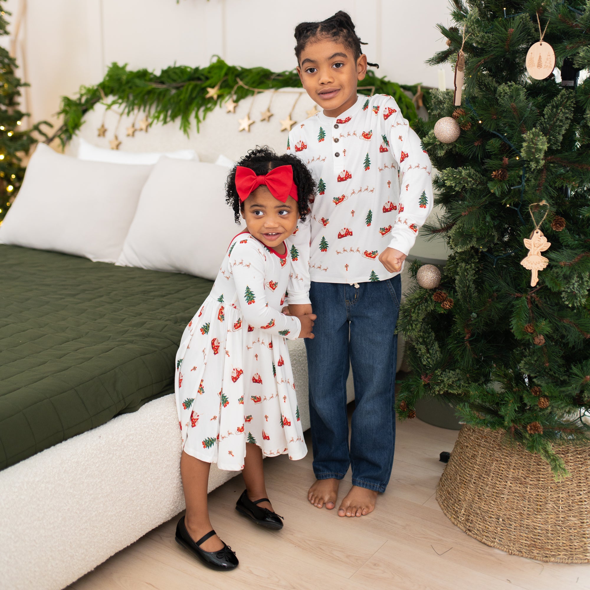 Siblings standing beside one another between a bed and christmas tree wearing the Long Sleeve Twirl Dress in Santa Sleigh and Long Sleeve Henley Tee in Santa Sleigh