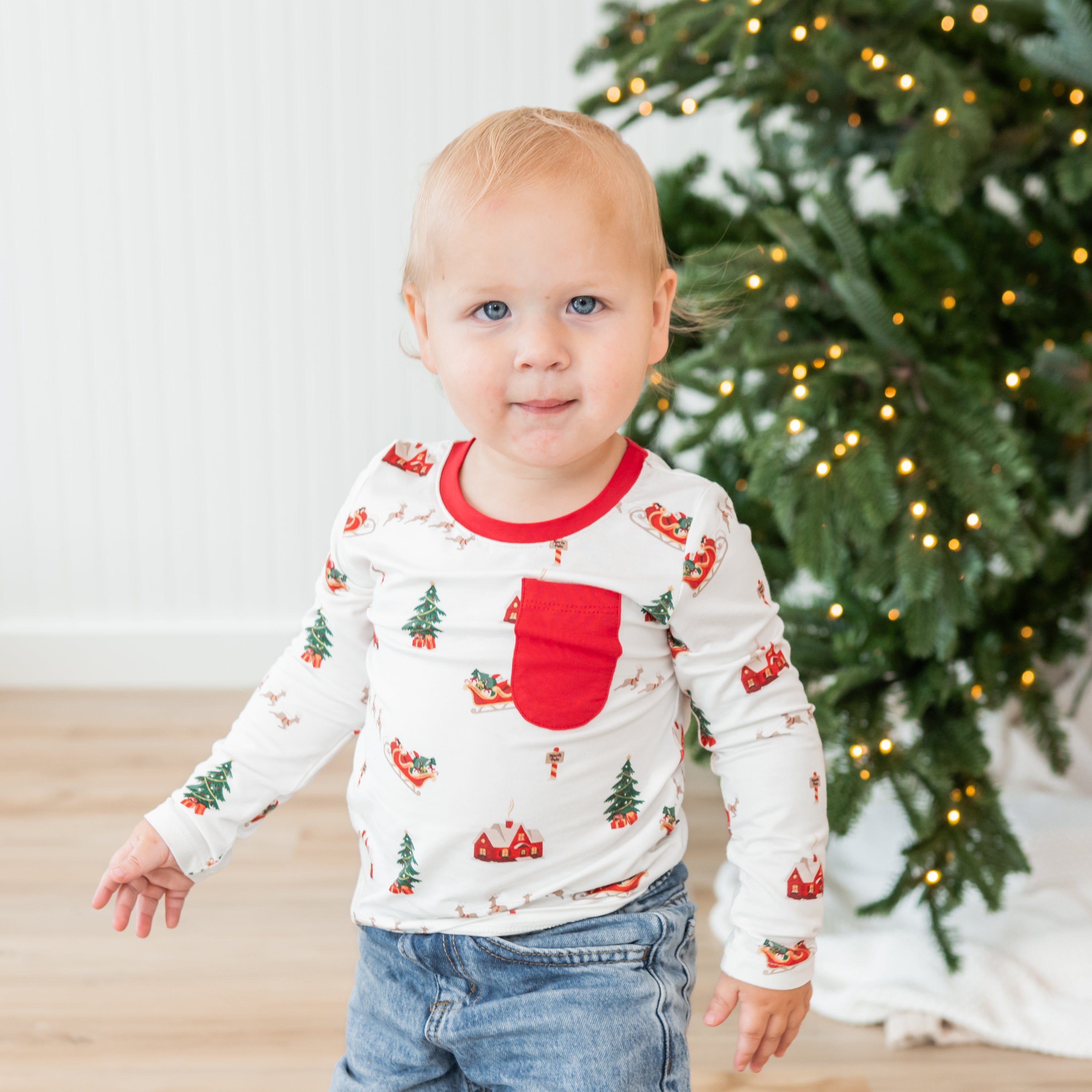 Young toddler standing in front of a Christmas tree wearing the Long Sleeve Toddler Crew Neck Tee in Santa Sleigh paired with light wash jeans
