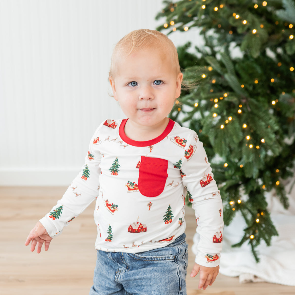 Young toddler standing in front of a Christmas tree wearing the Long Sleeve Toddler Crew Neck Tee in Santa Sleigh paired with light wash jeans