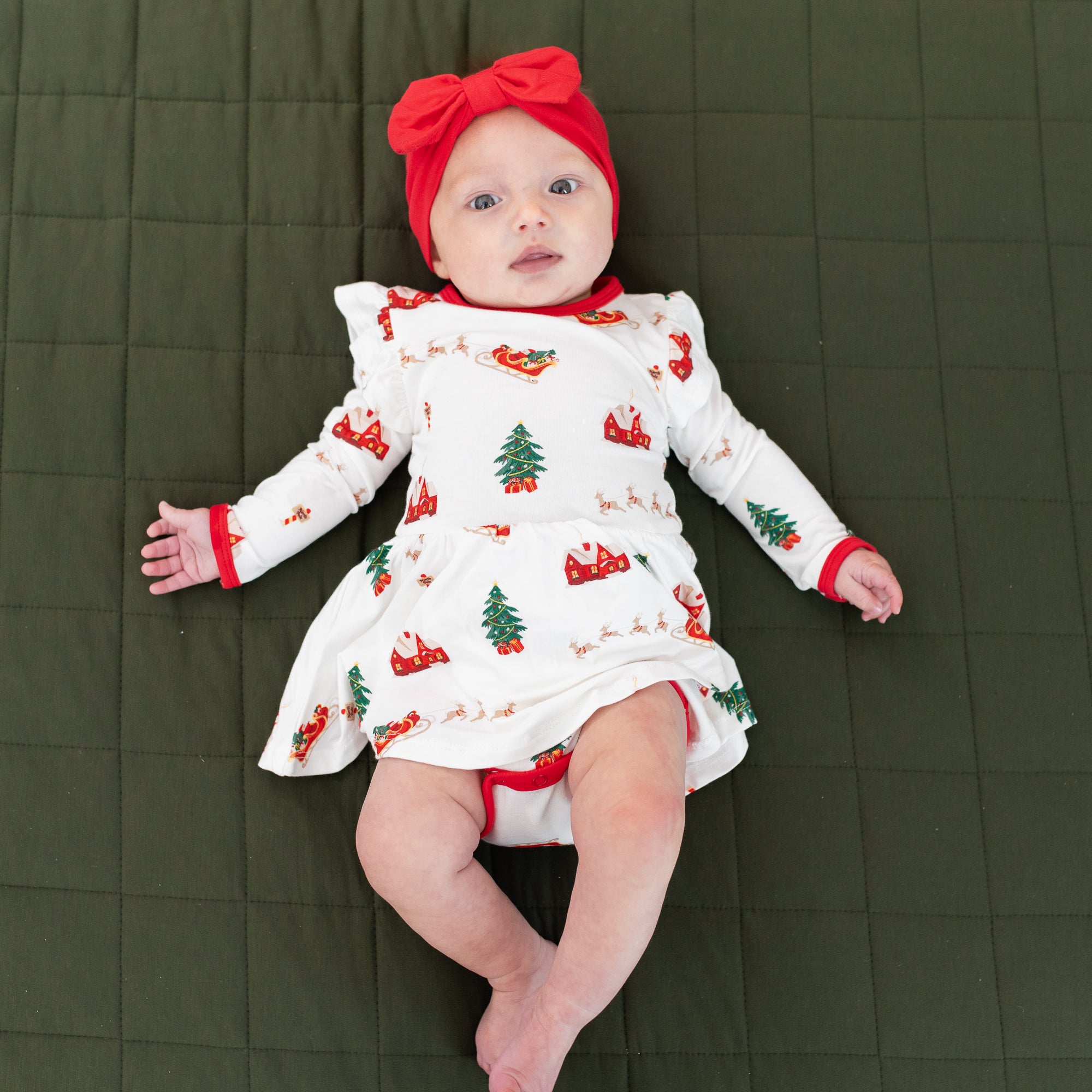 Young infant laying on a Fir blanket wearing the Long Sleeve Twirl Bodysuit Dress in Santa Sleigh and Cardinal bow headband