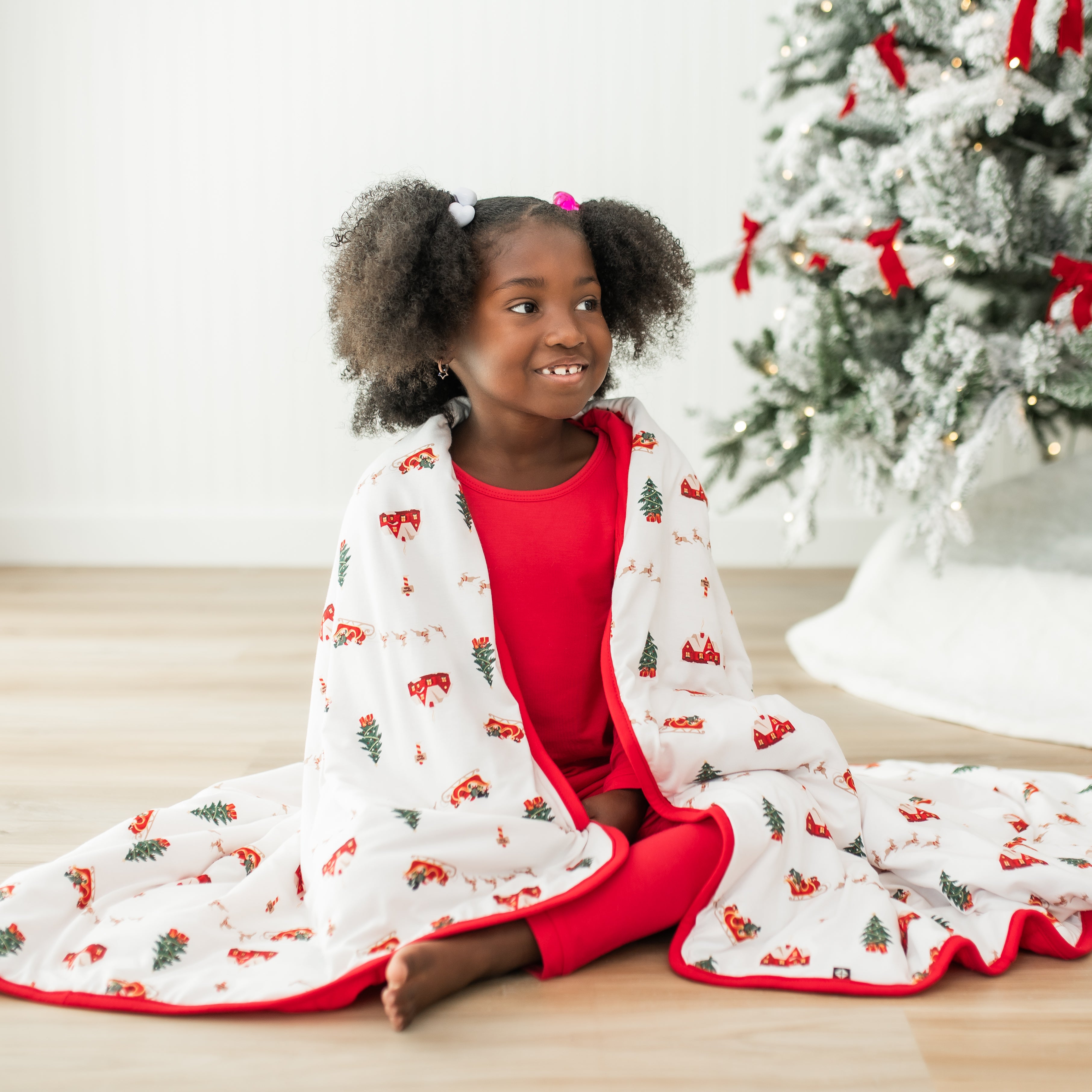 Smiling girl sitting on the floor wearing red pajamas with the Toddler Blanket in Santa Sleigh 1.0 over her shoulder