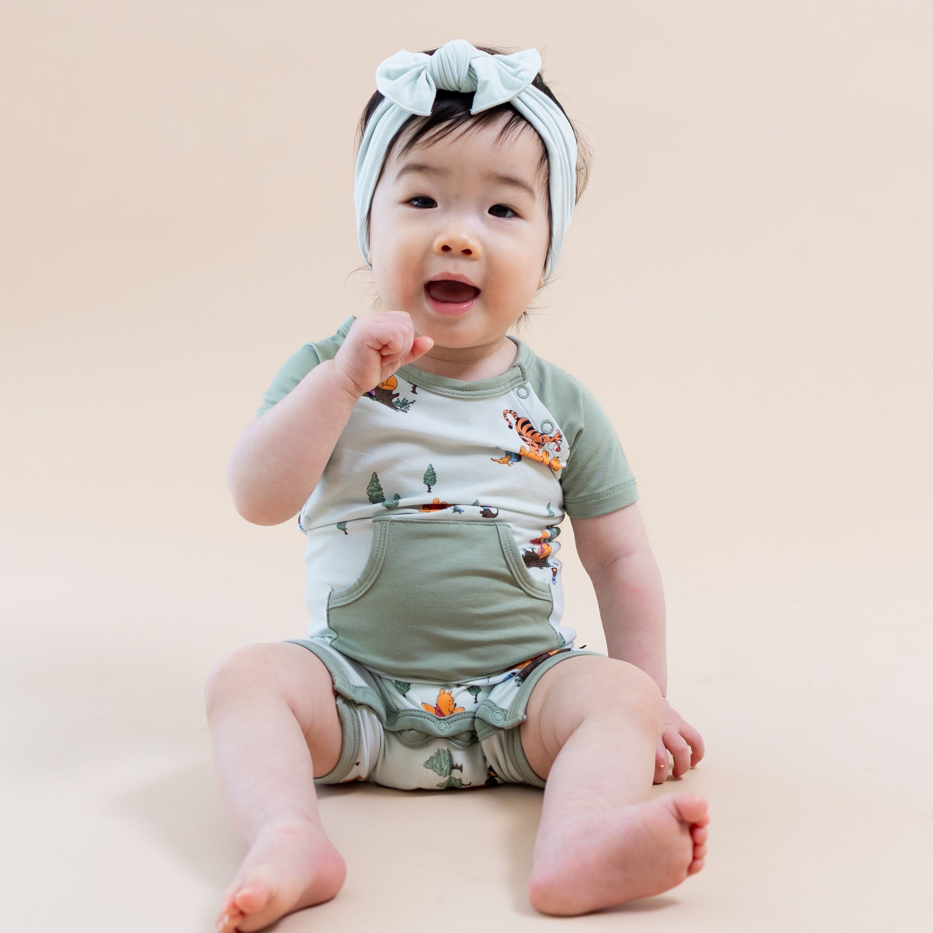 Young toddler sitting on the floor wearing the Shortall in Winnie the Pooh Hundred Acre Wood with light green bow