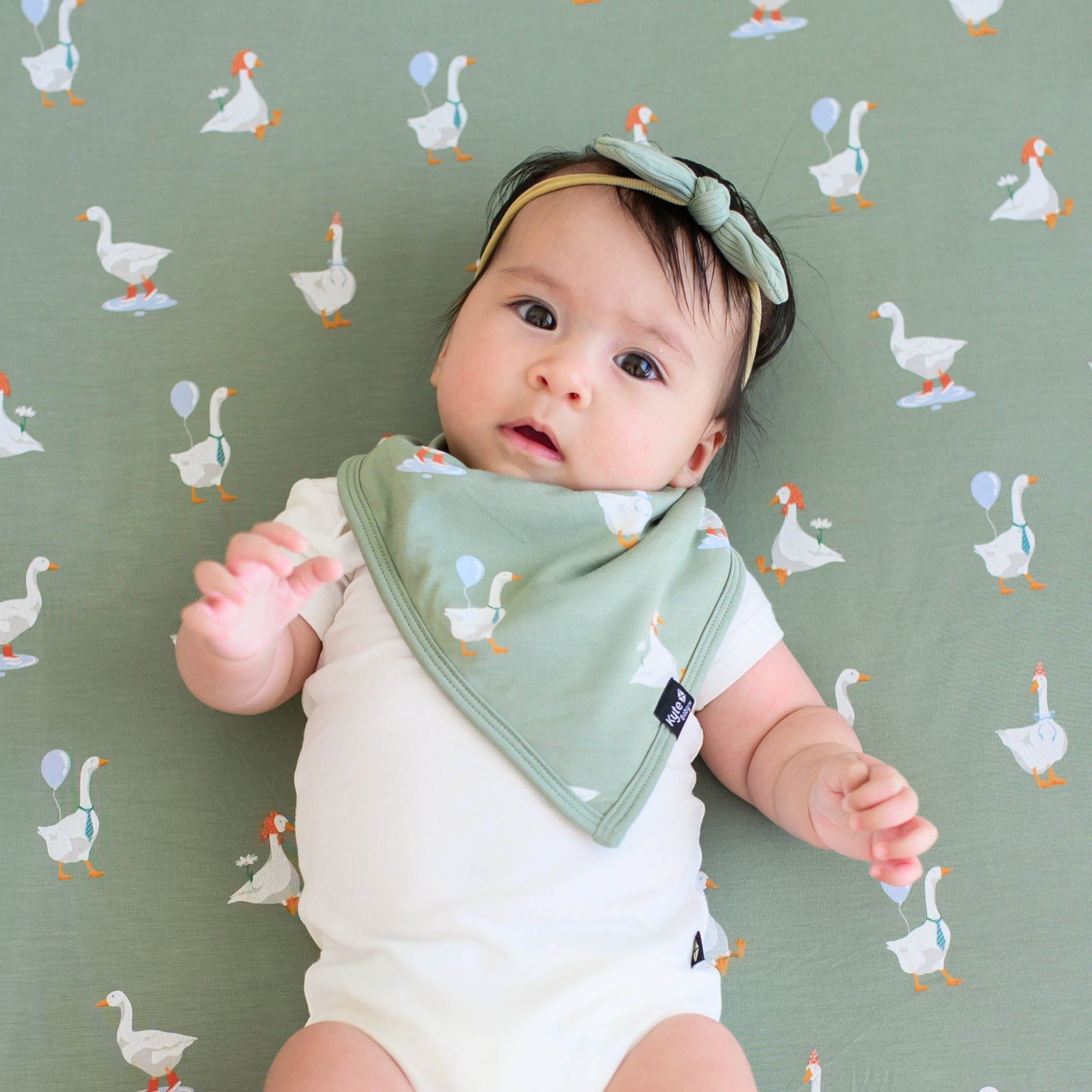 Infant laying on a Silly goose crib sheet wearing the Bib in Silly Goose, cloud bodysuit and ribbed nylon bow in Thyme