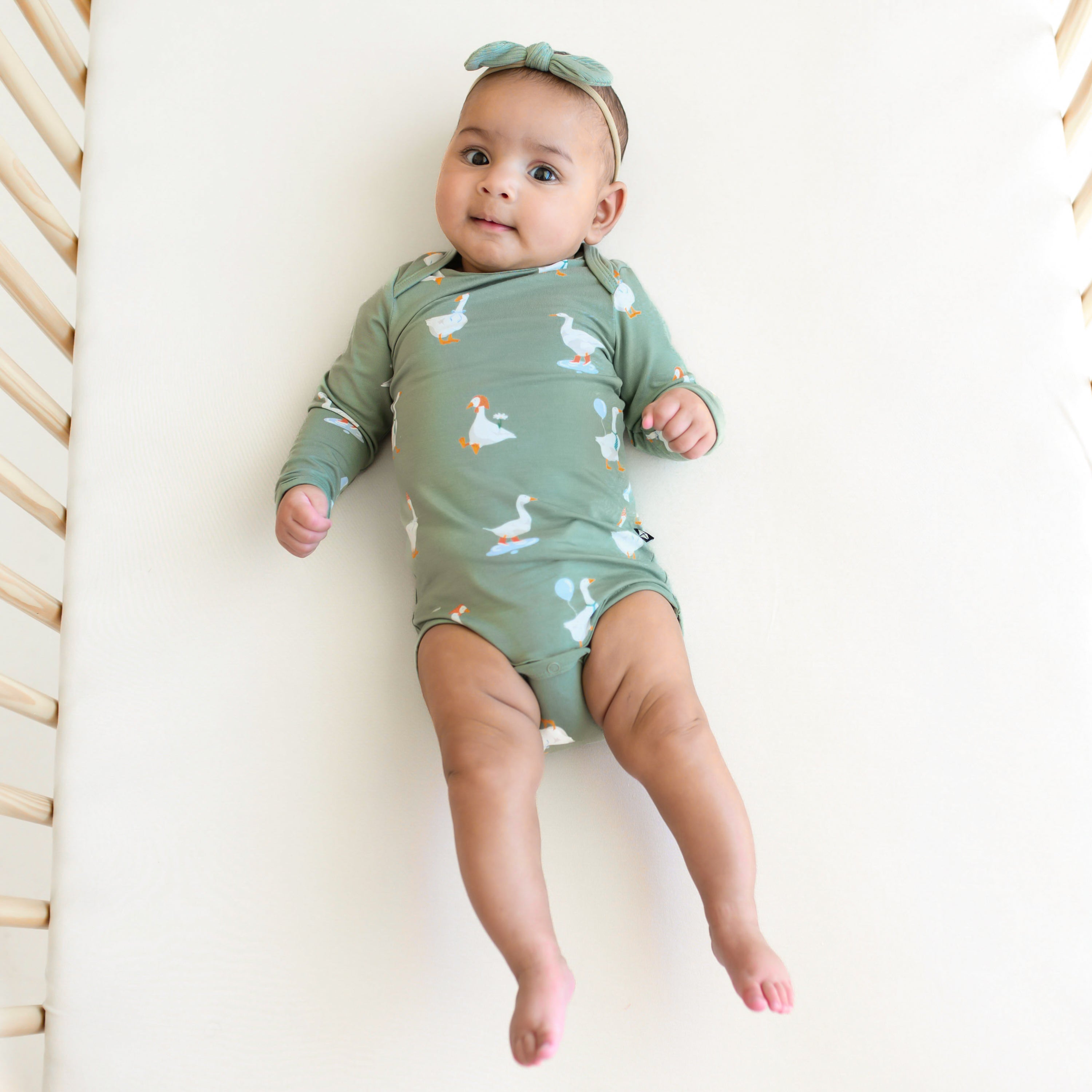 Infant girl laying in a crib on a neutral crib sheet wearing the Long Sleeve Bodysuit in Silly Goose and Ribbed Nylon Tiny Bow in Thyme