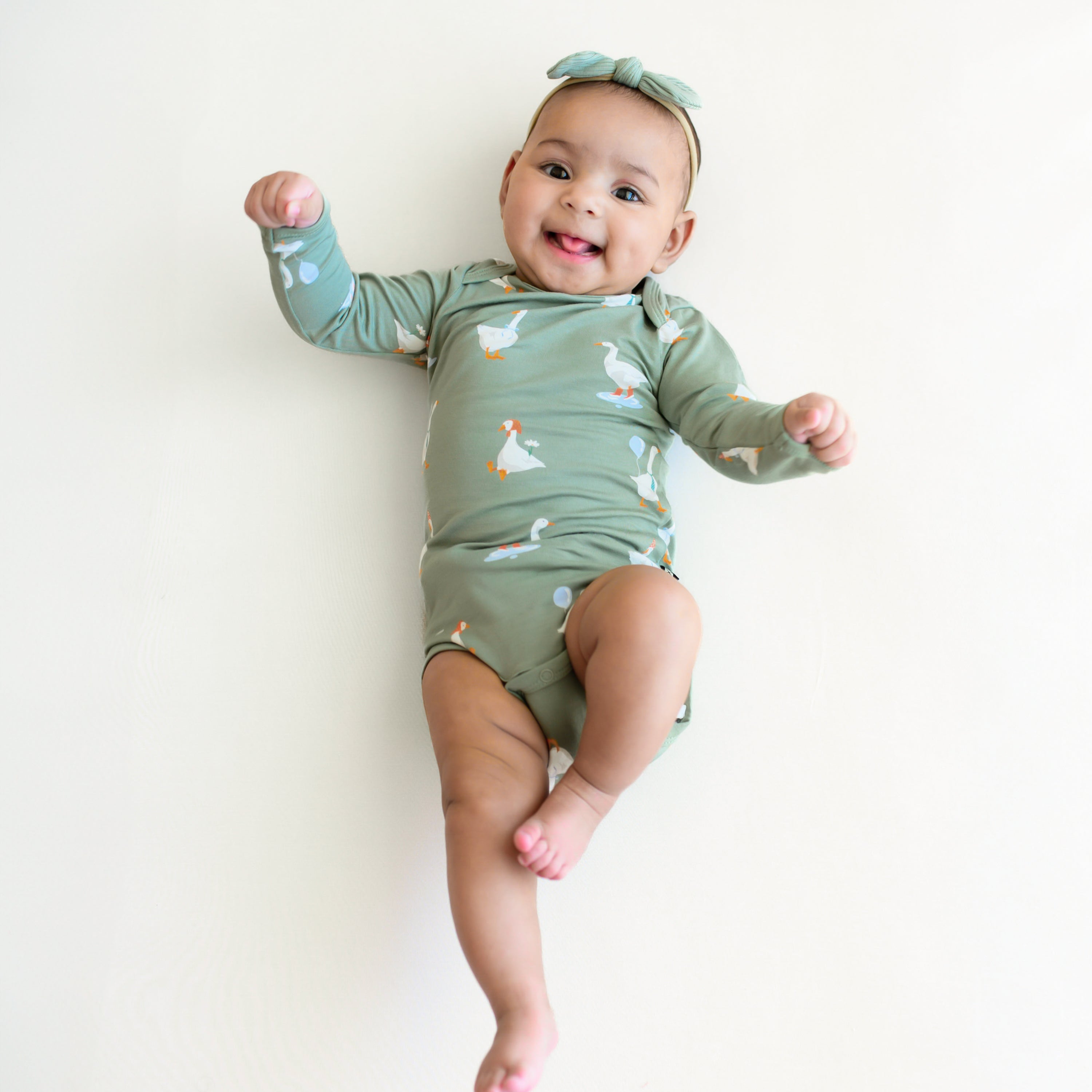 Infant laying on a blanket sticking her tongue out wearing the Long Sleeve Bodysuit in Silly Goose and Ribbed Nylon Tiny Bow in Thyme