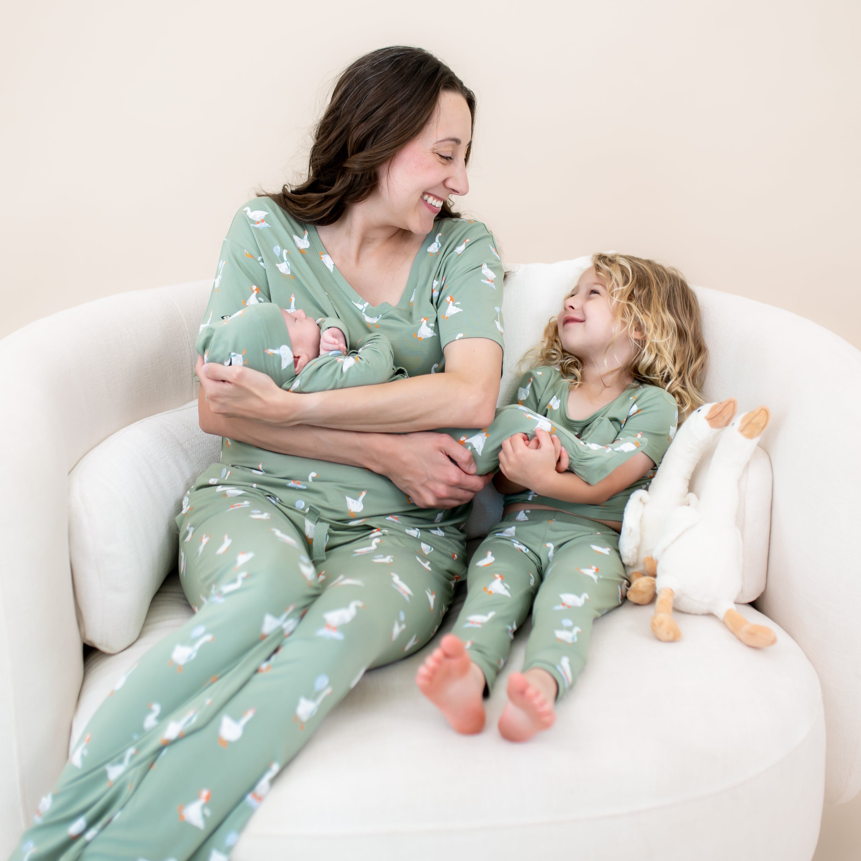 Family matching in Silly Goose sitting on a couch. Pictured is the women's v-neck, the take me home set with cap and the Short Sleeve with Pants Pajamas in Silly Goose