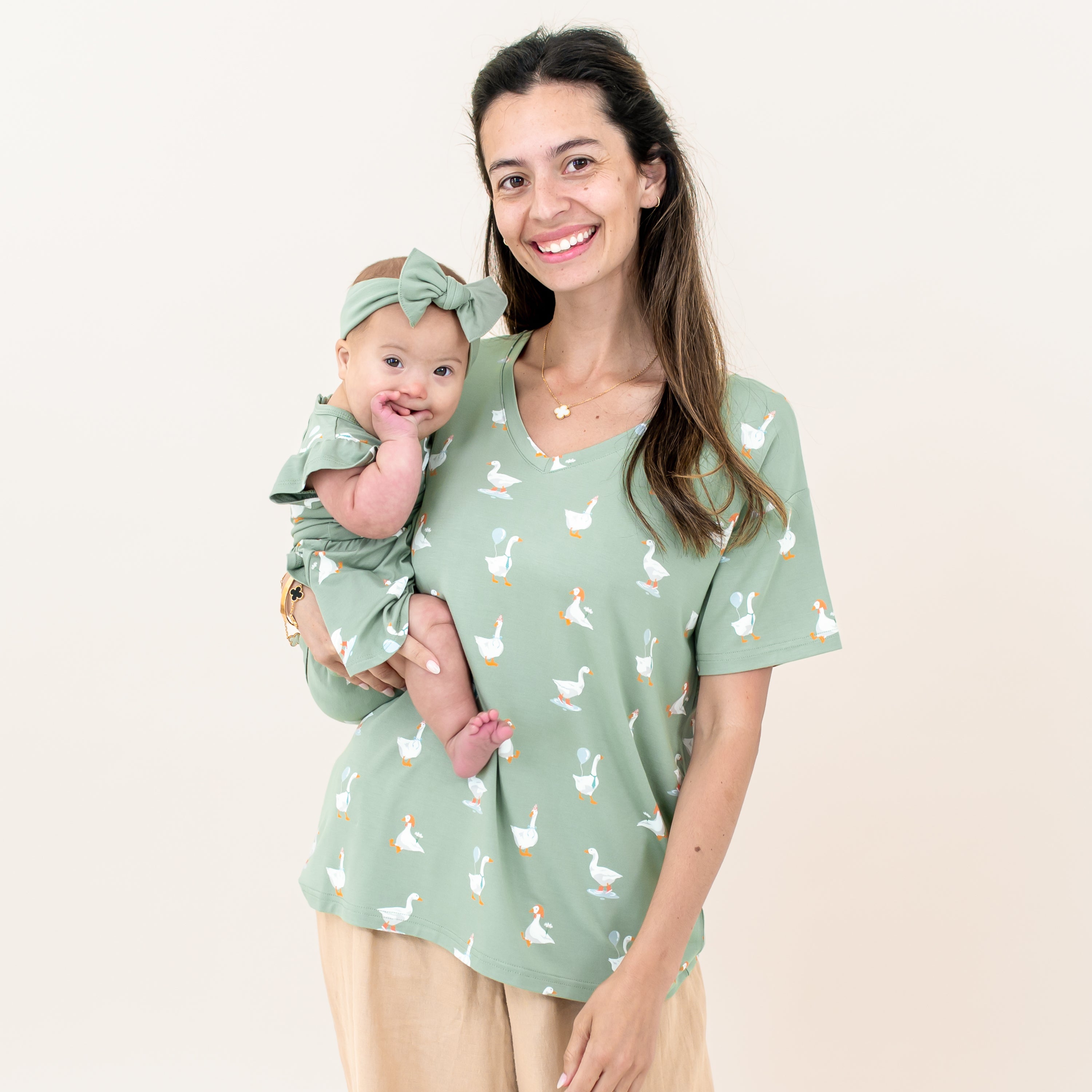 Smiling mother wearing the Women's Relaxed Fit V-Neck in Silly Goose with khaki colored pants holding her infant daughter who is matching with a Twirl Bodysuit dress and thyme knotted bow headband