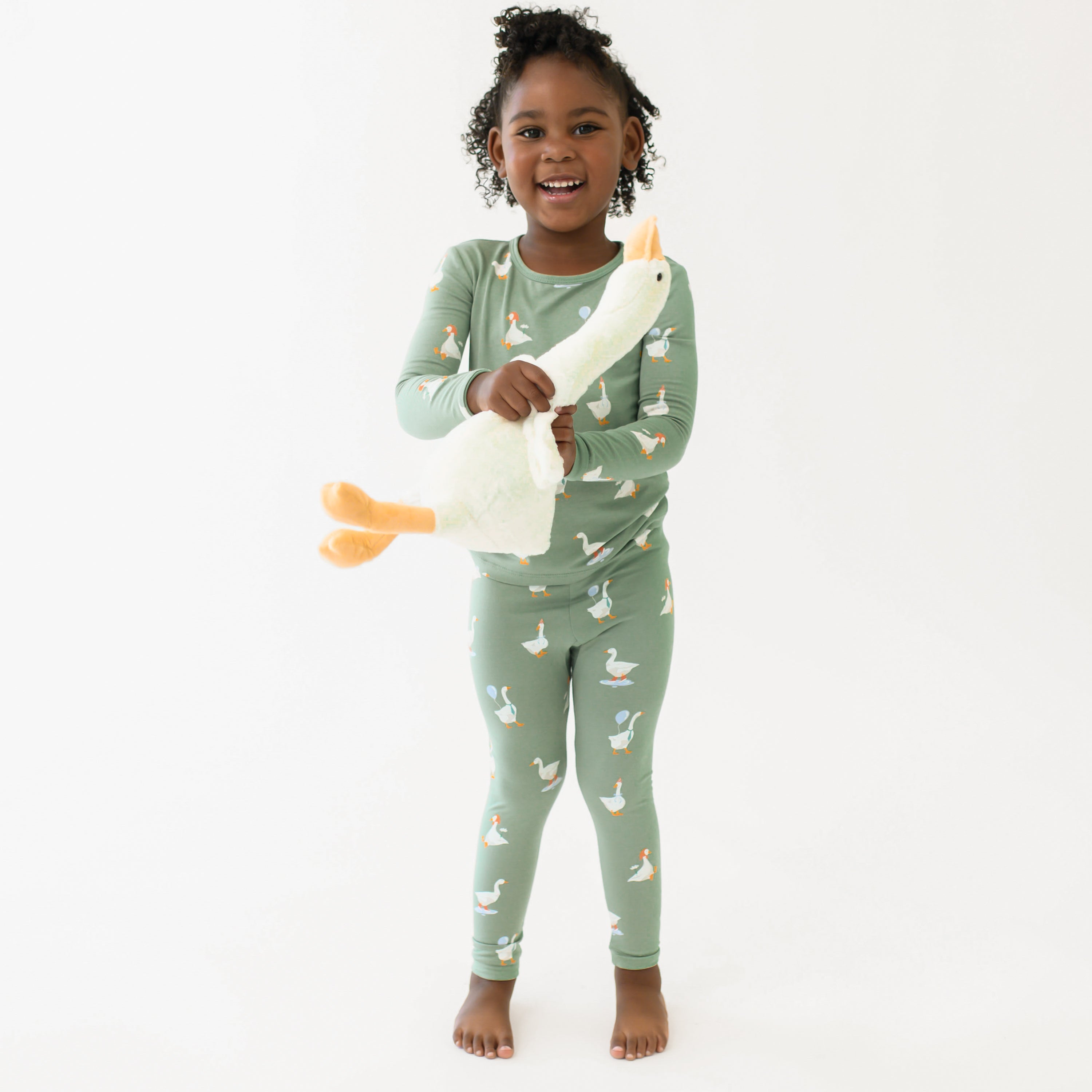 Smiling girl standing with a toy stuffed goose wearing the Long Sleeve Pajamas in Silly Goose in front of a white backdrop