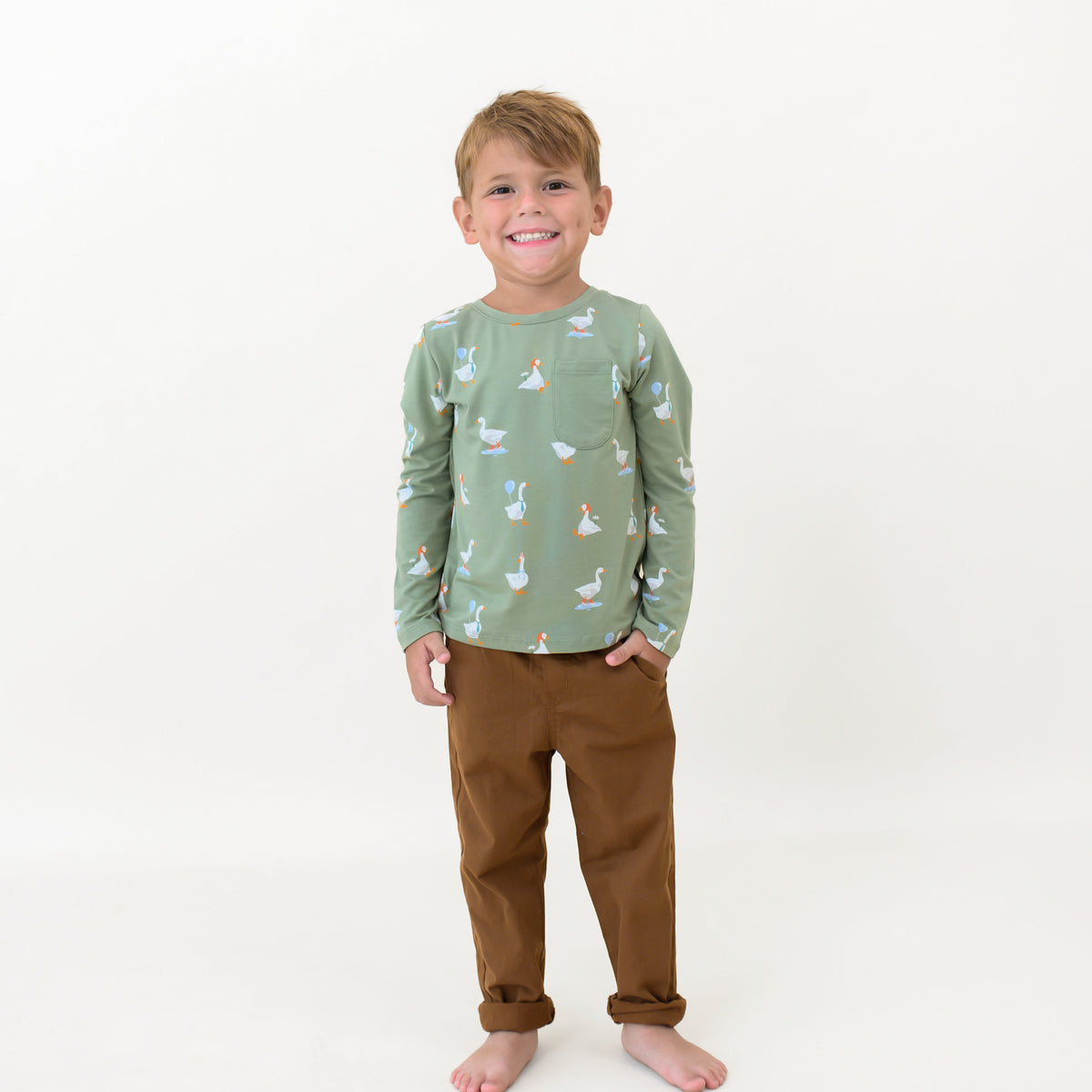 Smiling boy standing with one hand in his pant pocket wearing the Long Sleeve Toddler Crew Neck Tee in Silly Goose in front of white backdrop