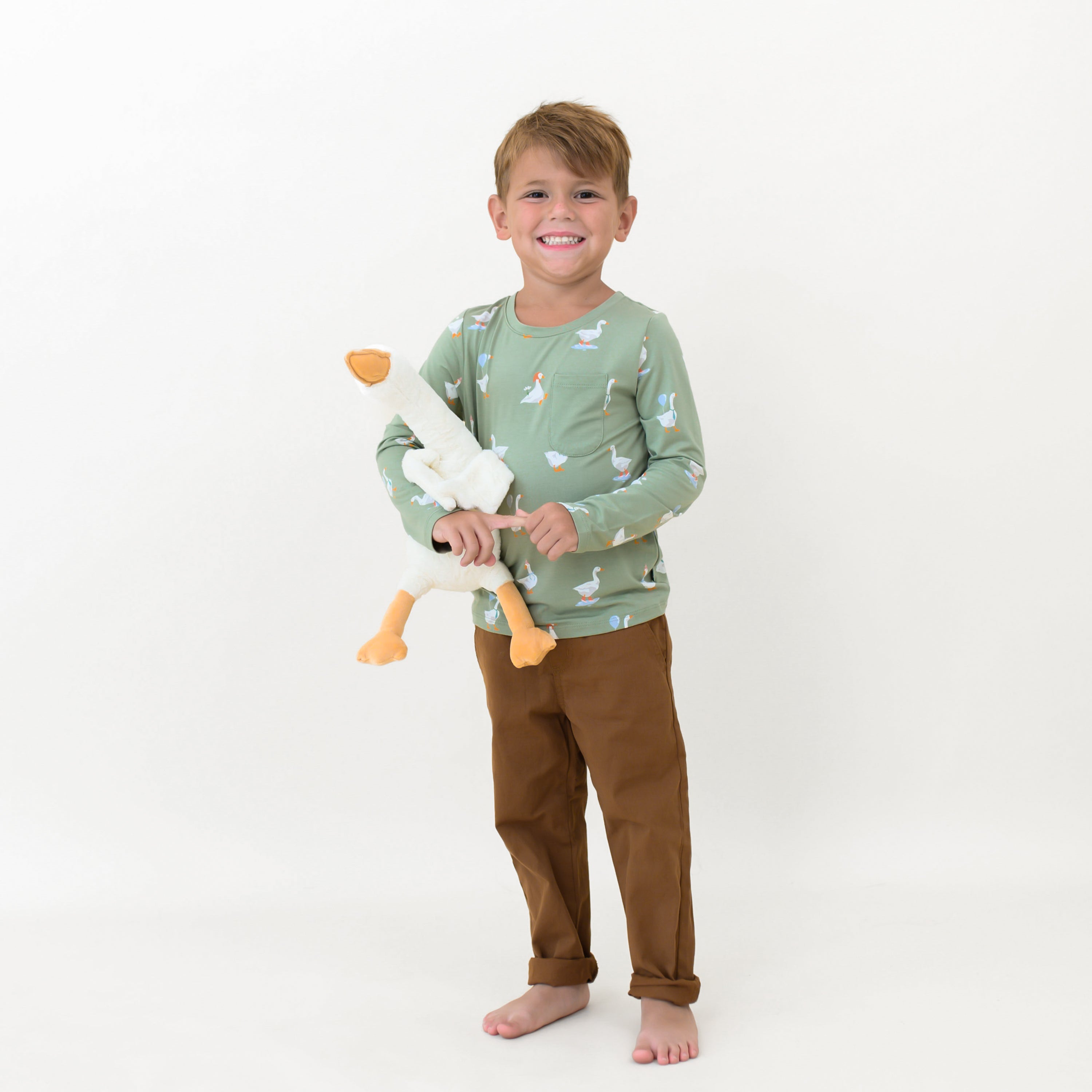 Smiling boy holding a toy stuffed goose wearing the Long Sleeve Toddler Crew Neck Tee in Silly Goose paired with brown pants in front of a white backdrop