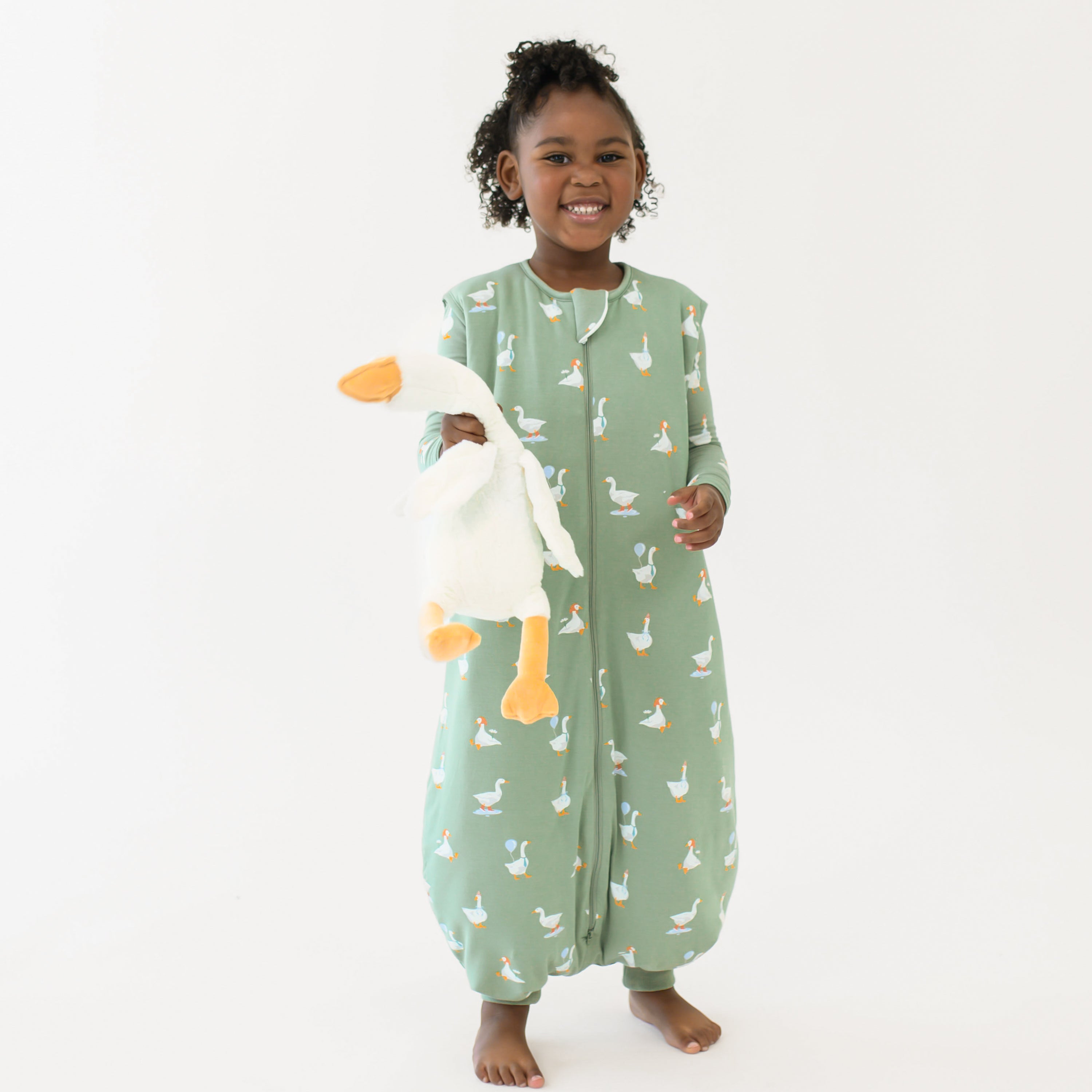 Smiling girl standing holding a toy stuffed goose wearing the Sleep Bag Walker in Silly Goose 1.0 and long sleeve toddler pajamas on underneath
