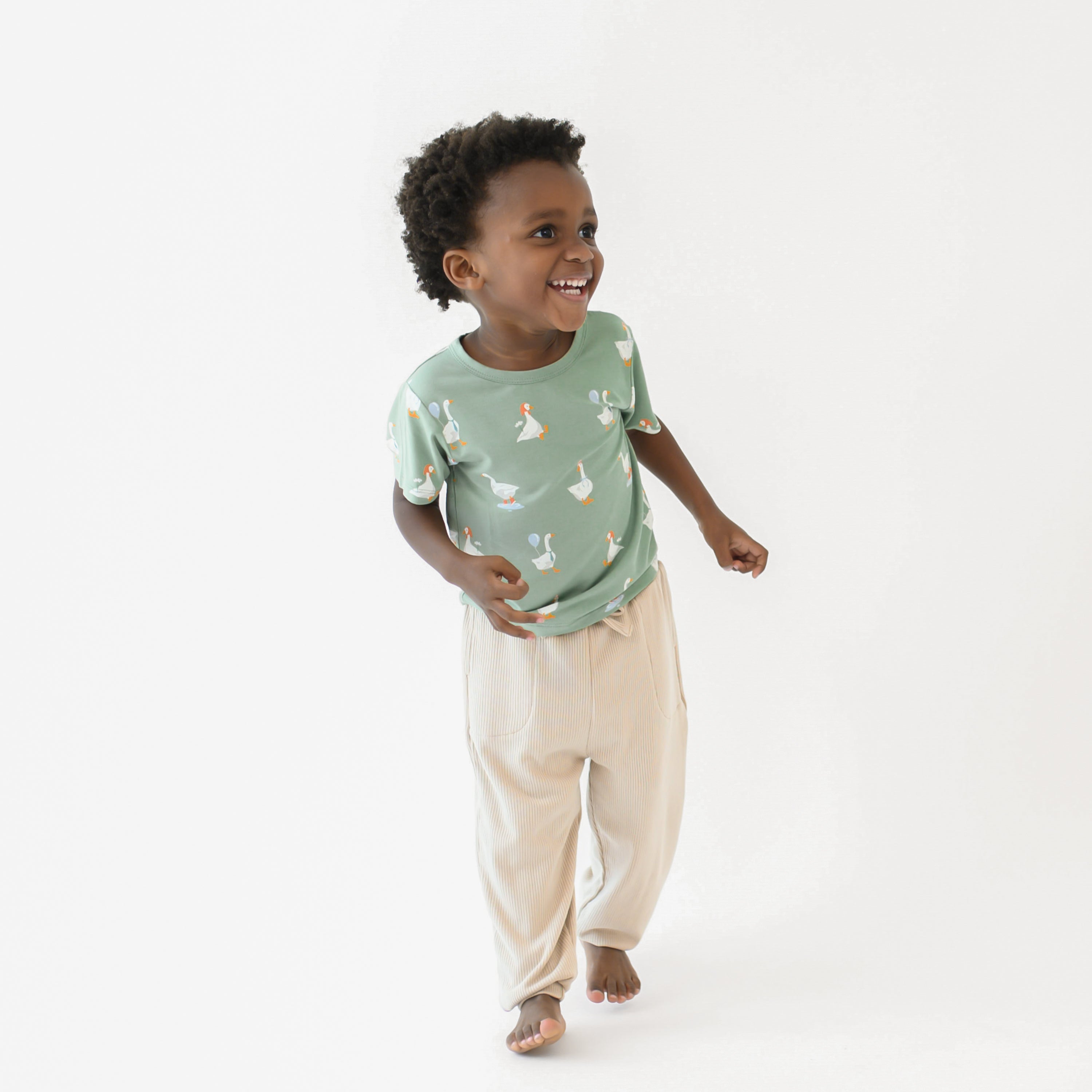 Young boy walking wearing the Toddler Basic Tee in Silly Goose and Bisque Ribbed Jogger pants in front of a white backdrp