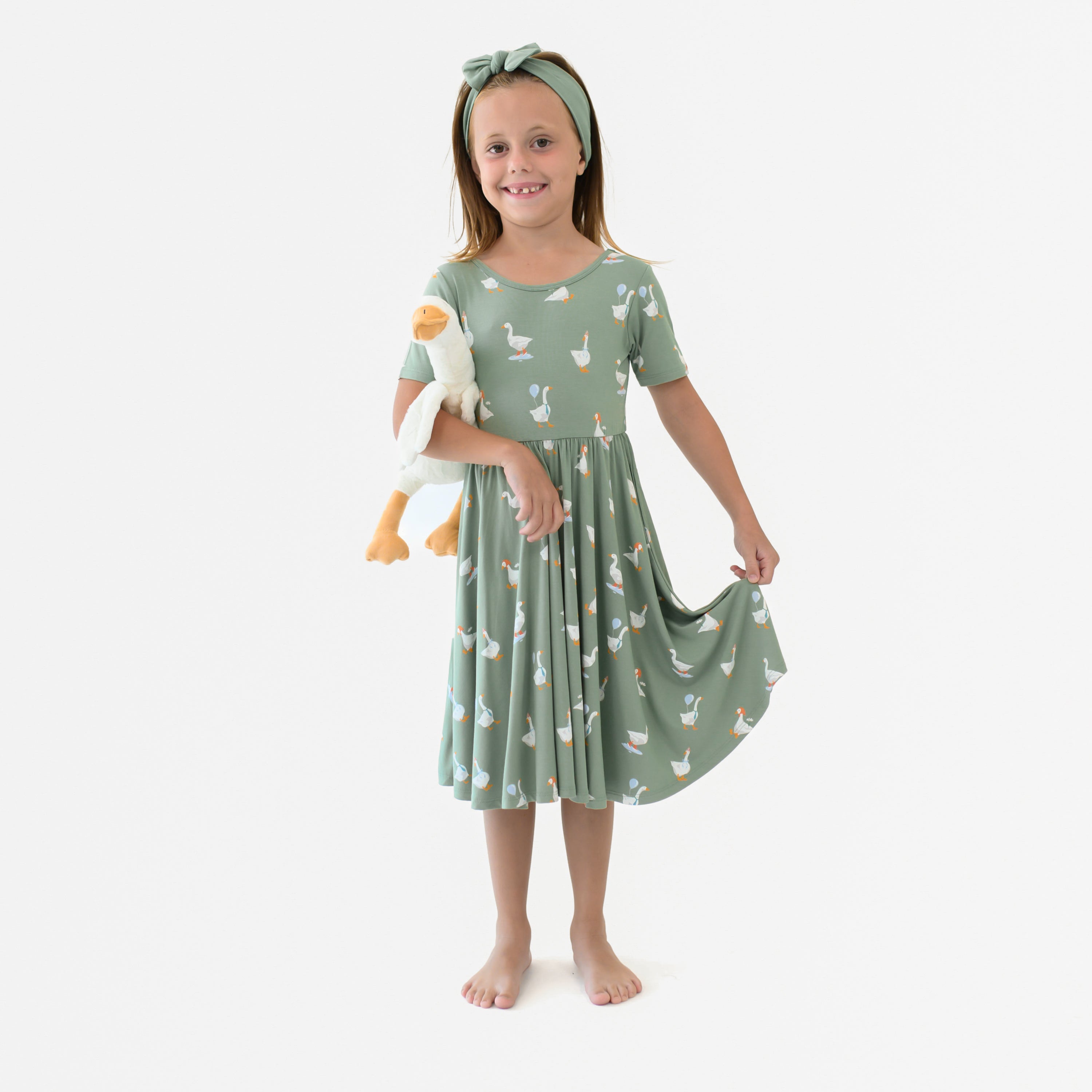 Young girl model standing holding the bottom of the hem of the Twirl Dress in Silly Goose holding a stuffed toy goose
