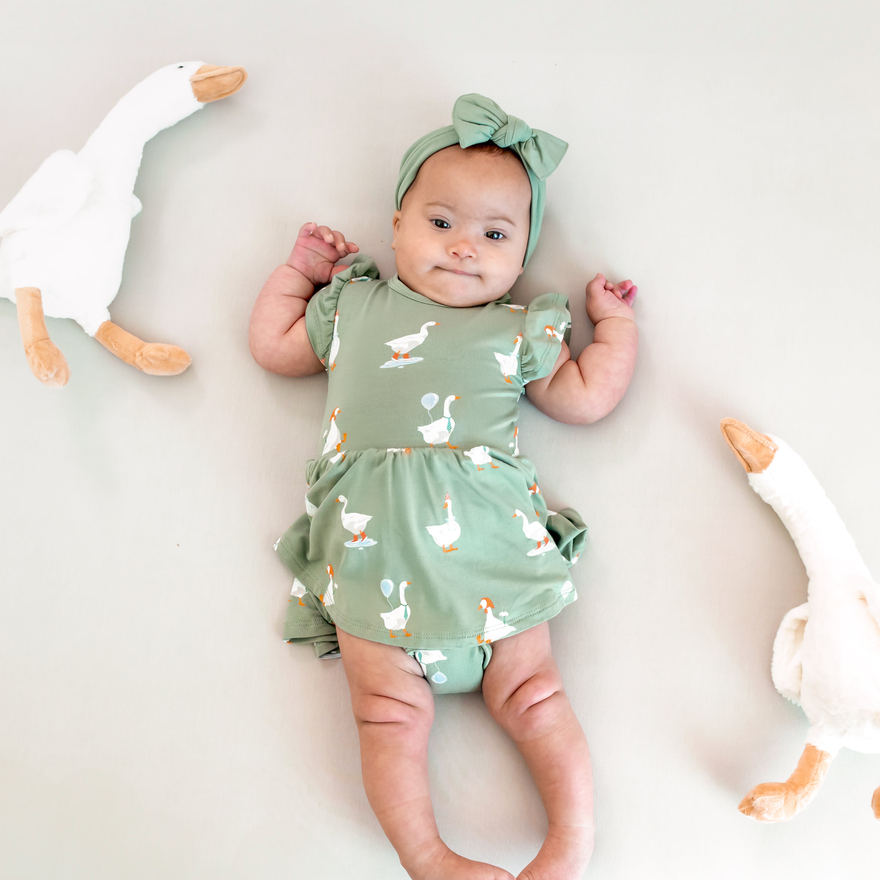 Infant laying on a light neutral background with two toy stuffed geese around her wearing the Twirl Bodysuit Dress in Silly Goose and knotted bow headband in Thyme