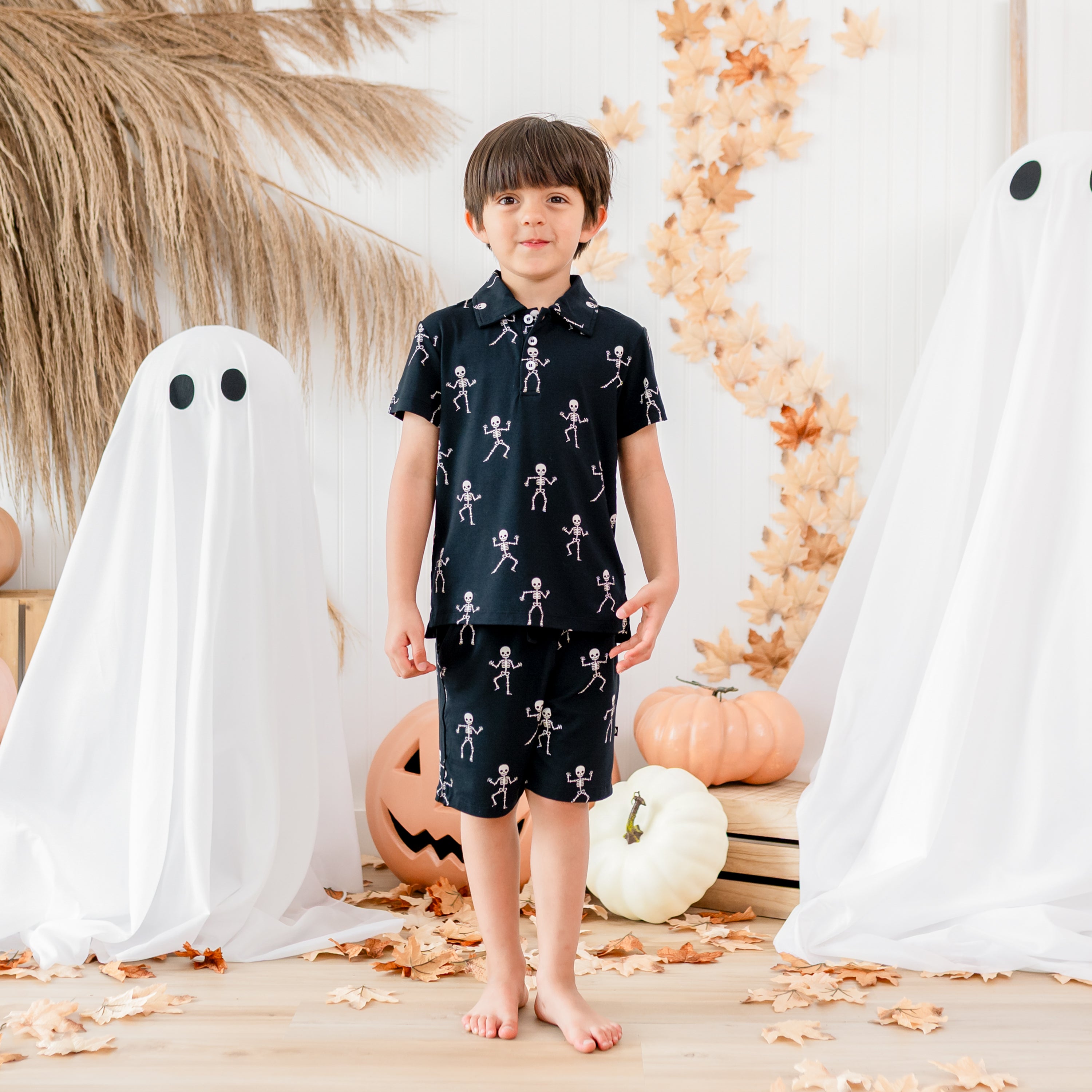 Boy model standing in front of a fall backdrop of sheet ghosts, pumpkins and leaves wearing the Toddler Short Sleeve Polo in Skeleton paired with Skeleton drawstring shorts