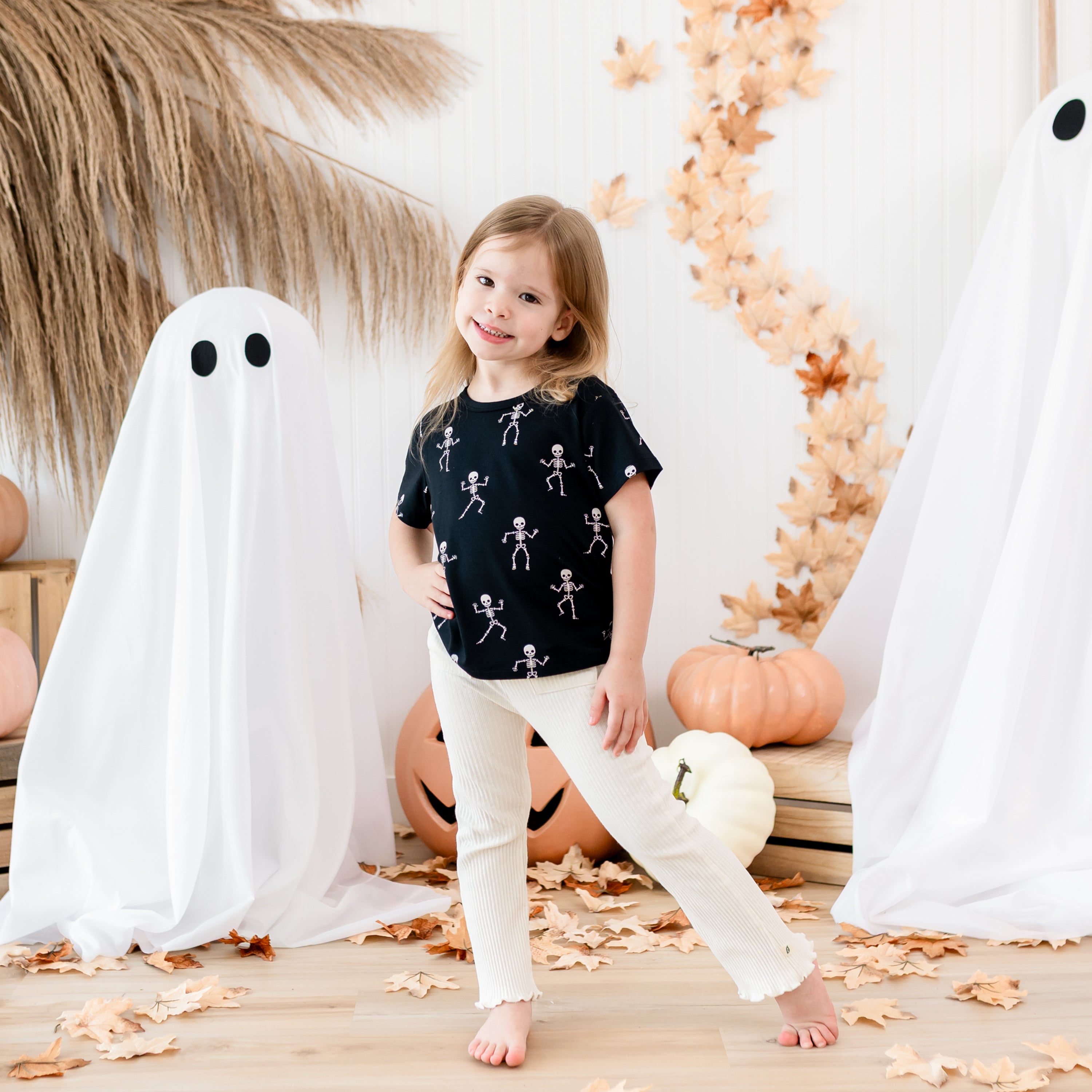 Smiling girl standing with one leg pointed outwards modeling the Toddler Basic Tee in Skeleton paired with light colored ribbed pants in front of halloween backdrop