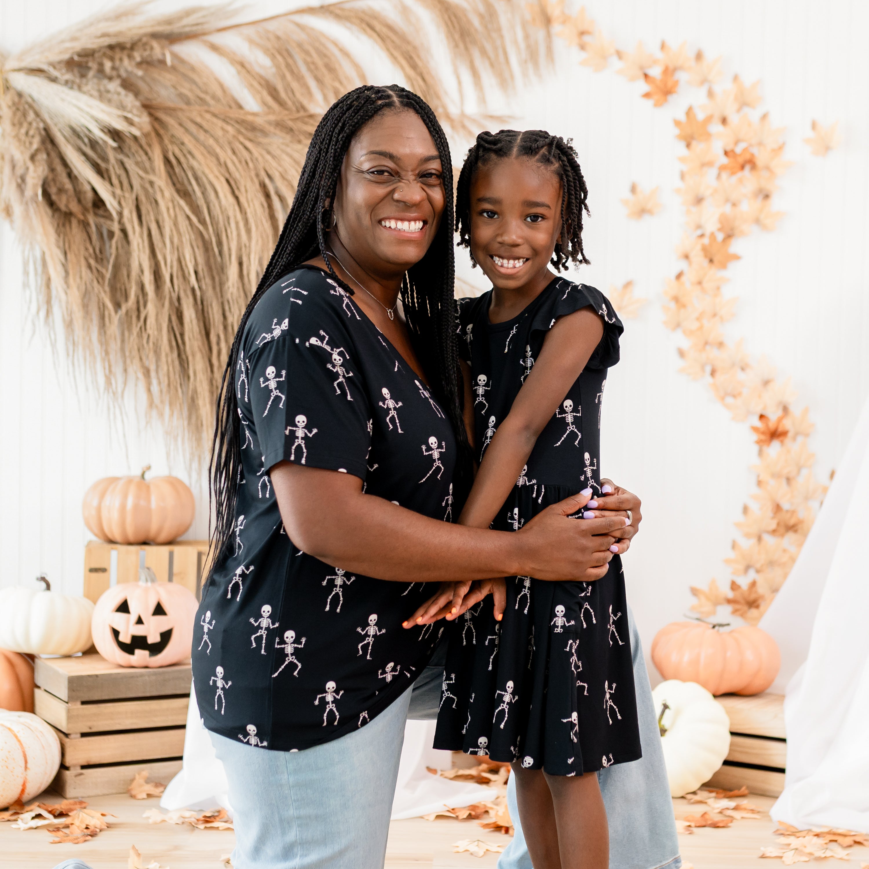 Mother and daughter matching in Skeleton. Mother is kneeling wearing the Women's Relaxed Fit V-Neck and jeans with her arms wrapped around her daughter who is wearing the pocket dress. Both are in front off a rustic fall inspired, halloween back drop