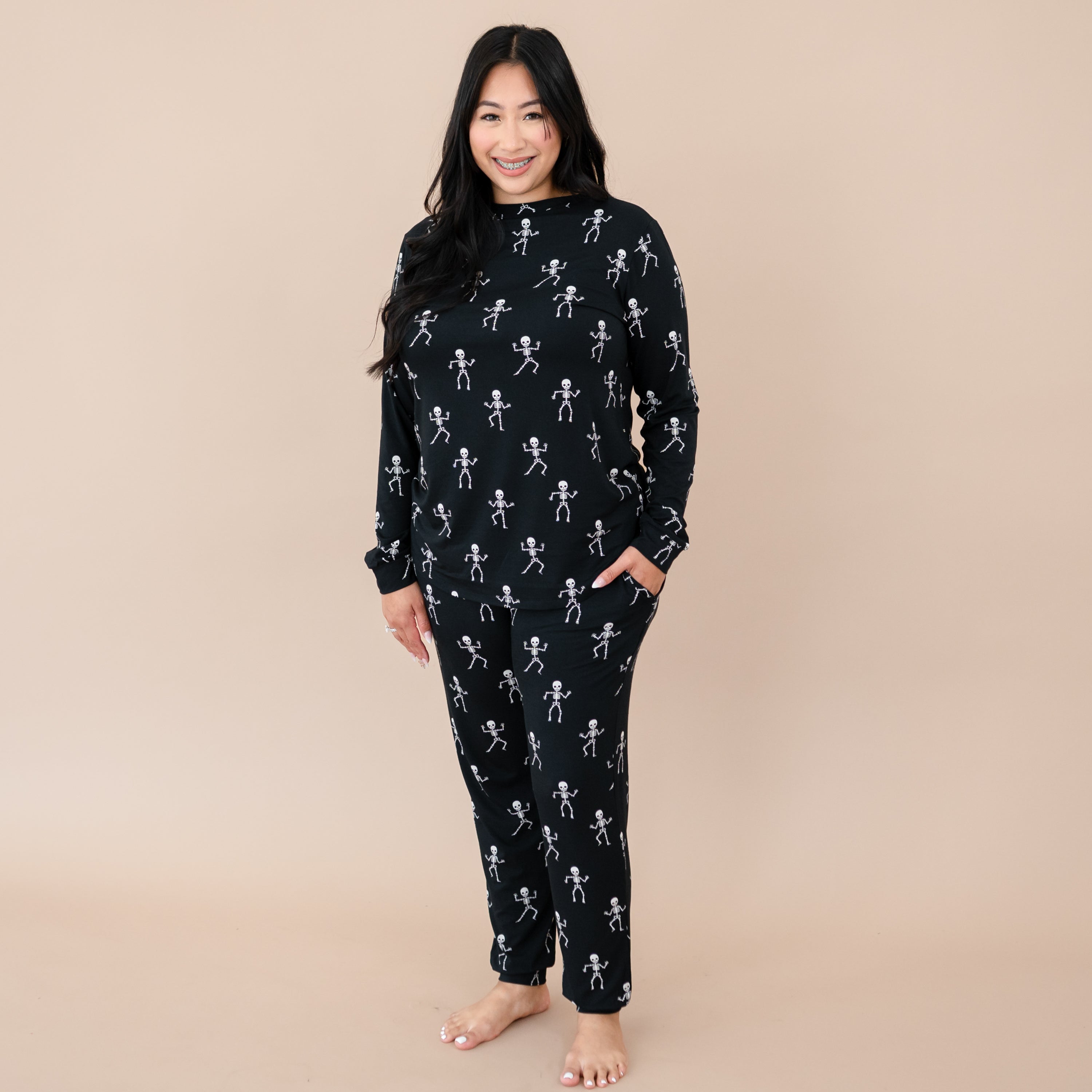 Smiling female wearing the Women's Jogger Pajama Set in Skeleton with one hand in the pants pocket in front of a taupe back drop