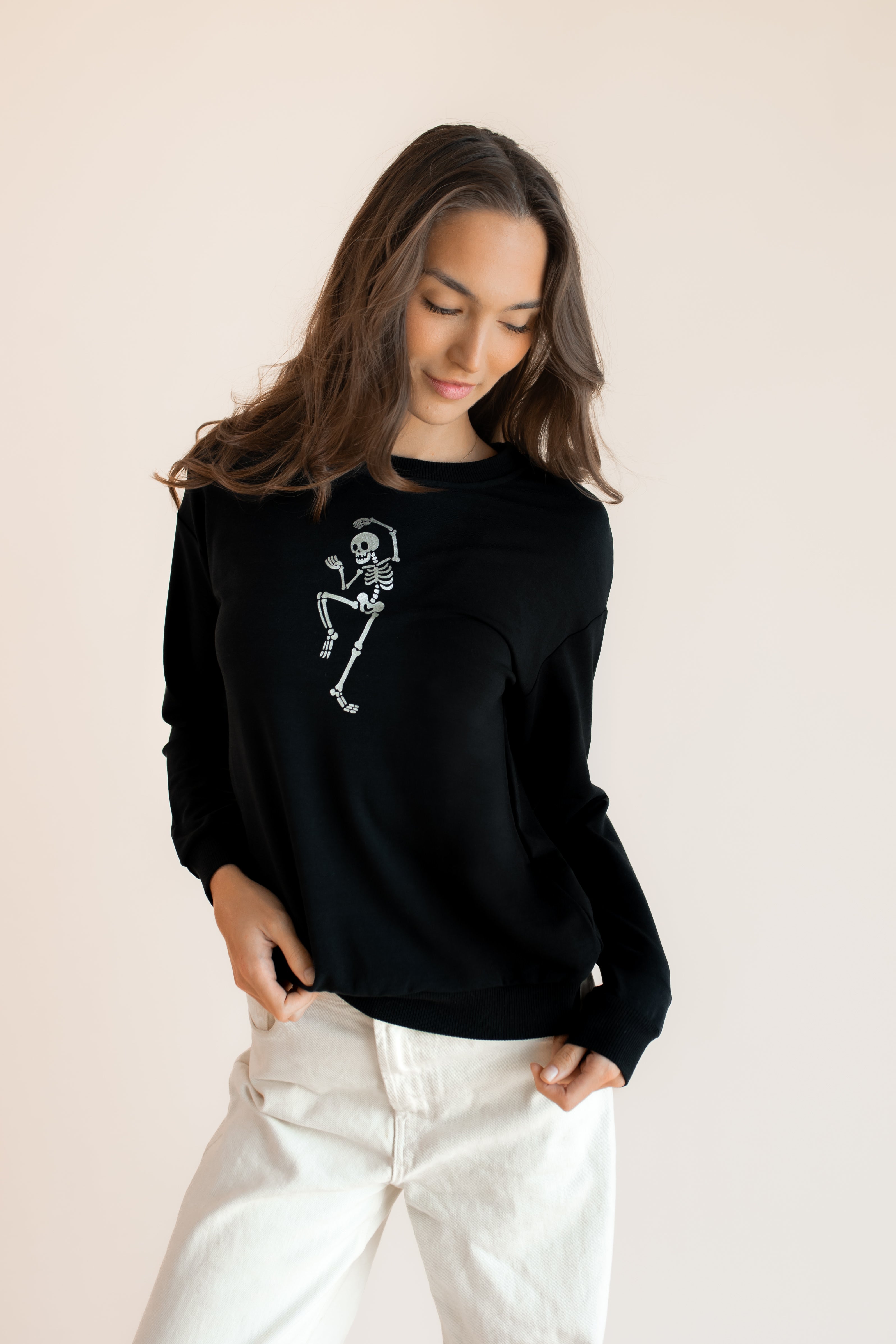 Modal wearing Black Women's Sweatshirt with Skeleton print in the center over white pants on a beige background