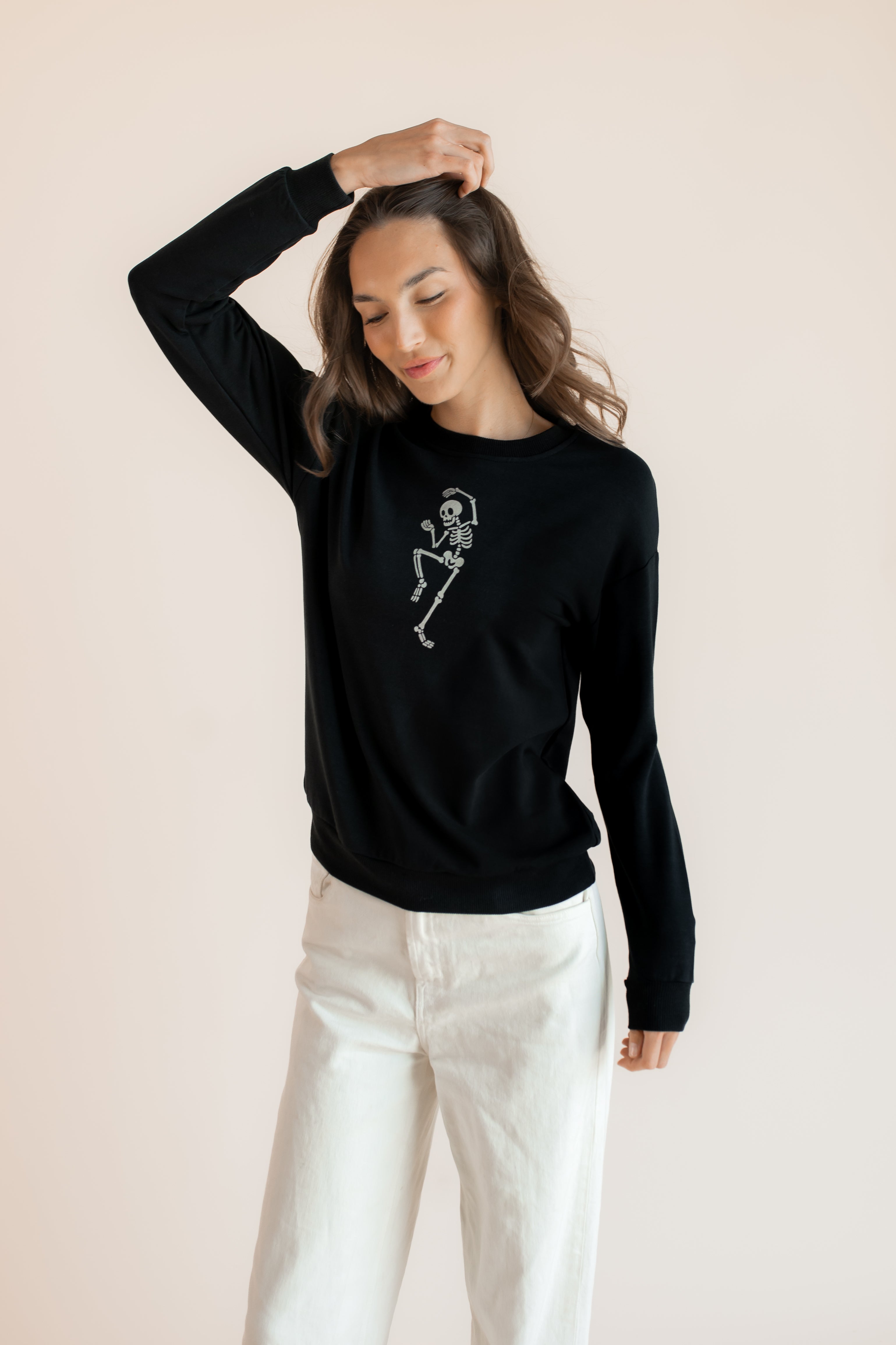 Modal posing in Black Women's Sweatshirt with Skeleton print in the center over white pants on a beige background