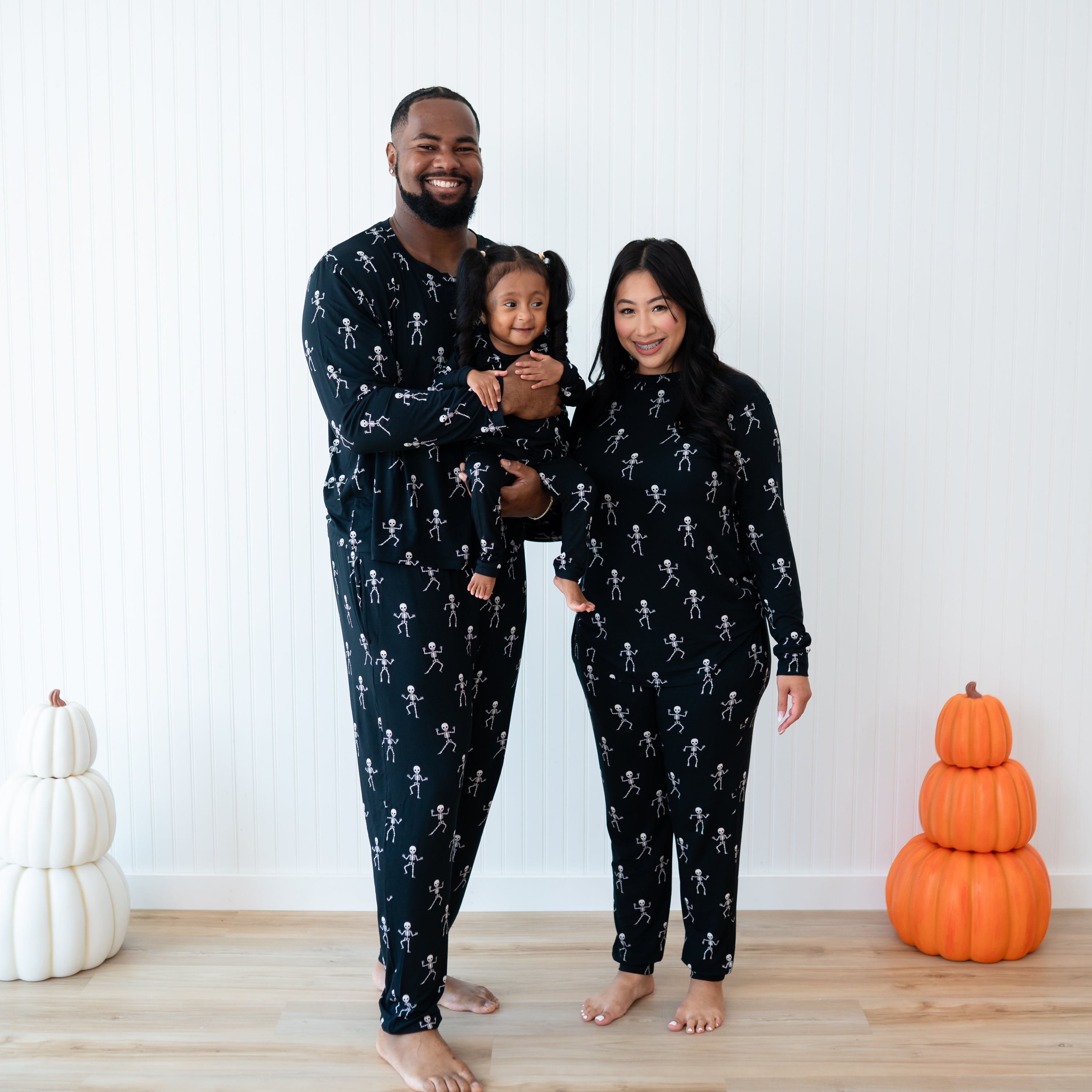 Family matching in Skeleton in front of a white wall and between stacked pumpkins. Father wearing the Men's jogger set is holding his daughter wearing the long sleeve toddler pajamas. Mother is standing beside them wearing the Women's Jogger Pajama Set
