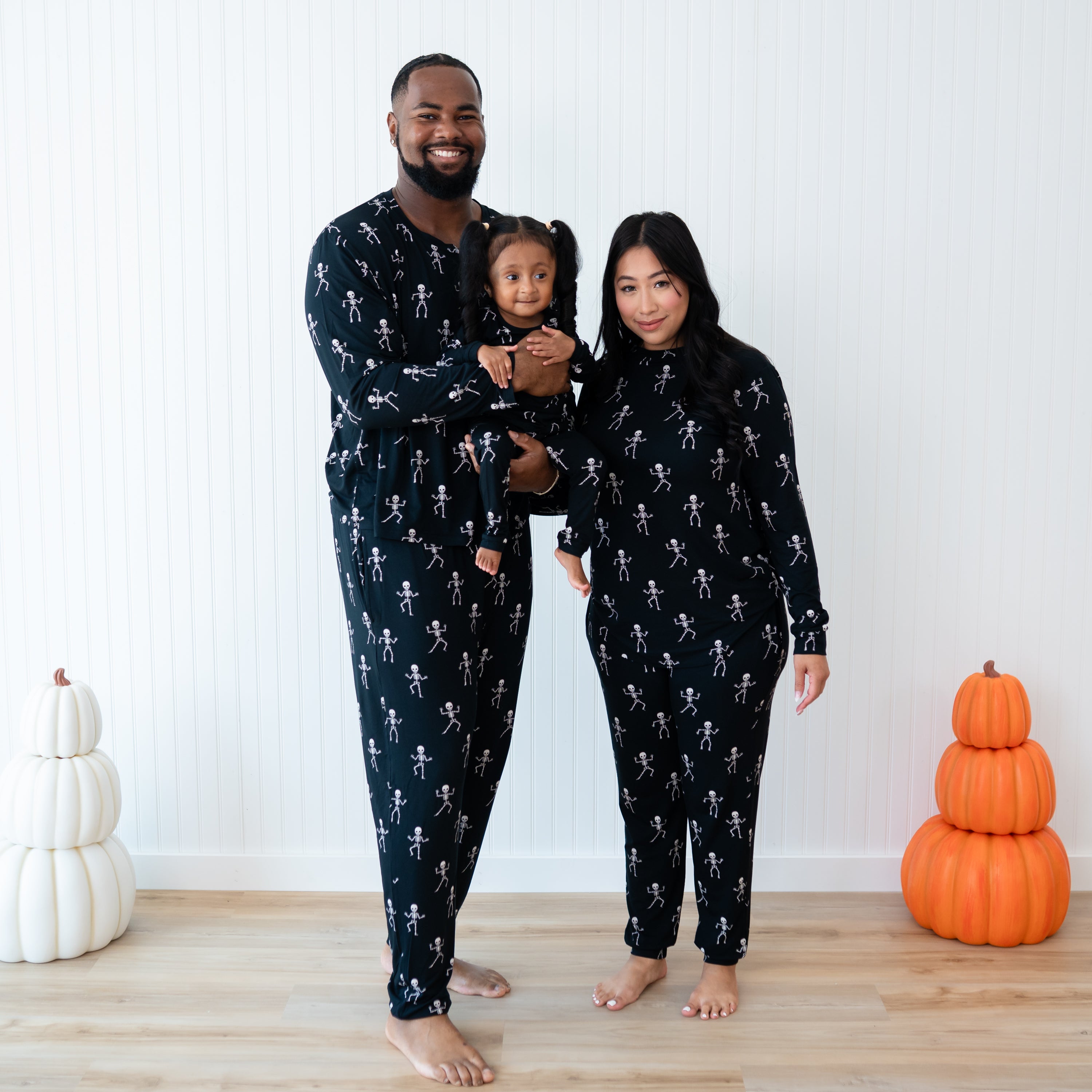 Family matching in Skeleton. Father wearing the Men's Jogger Set holding the daughter wearing the Toddler long sleeve pajama set. Mother is standing beside them wearing the Women's Jogger set