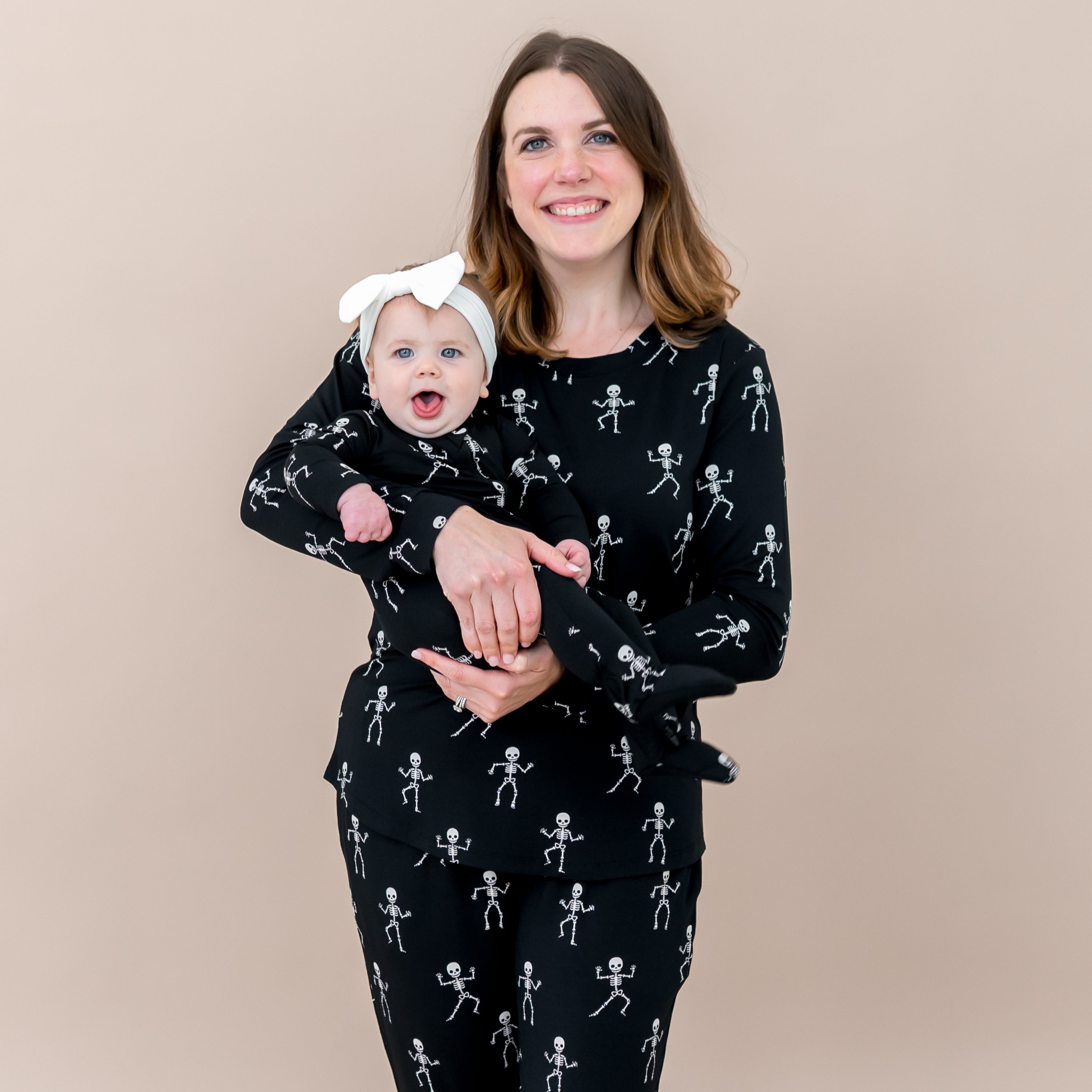 Mother and daughter matching in Skeleton. Smiling mother wearing the Women's Jogger Pajama Set holding her infant daughter who is wearing the zippered footie in front of a taupe backdrop