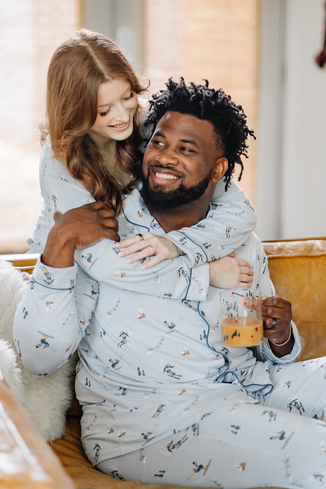 Male sitting on the couch wearing the Men's Long Sleeve Pajama Set in Ski with his wife standing behind him with her arms around her shoulders matching in women's ski pajama set