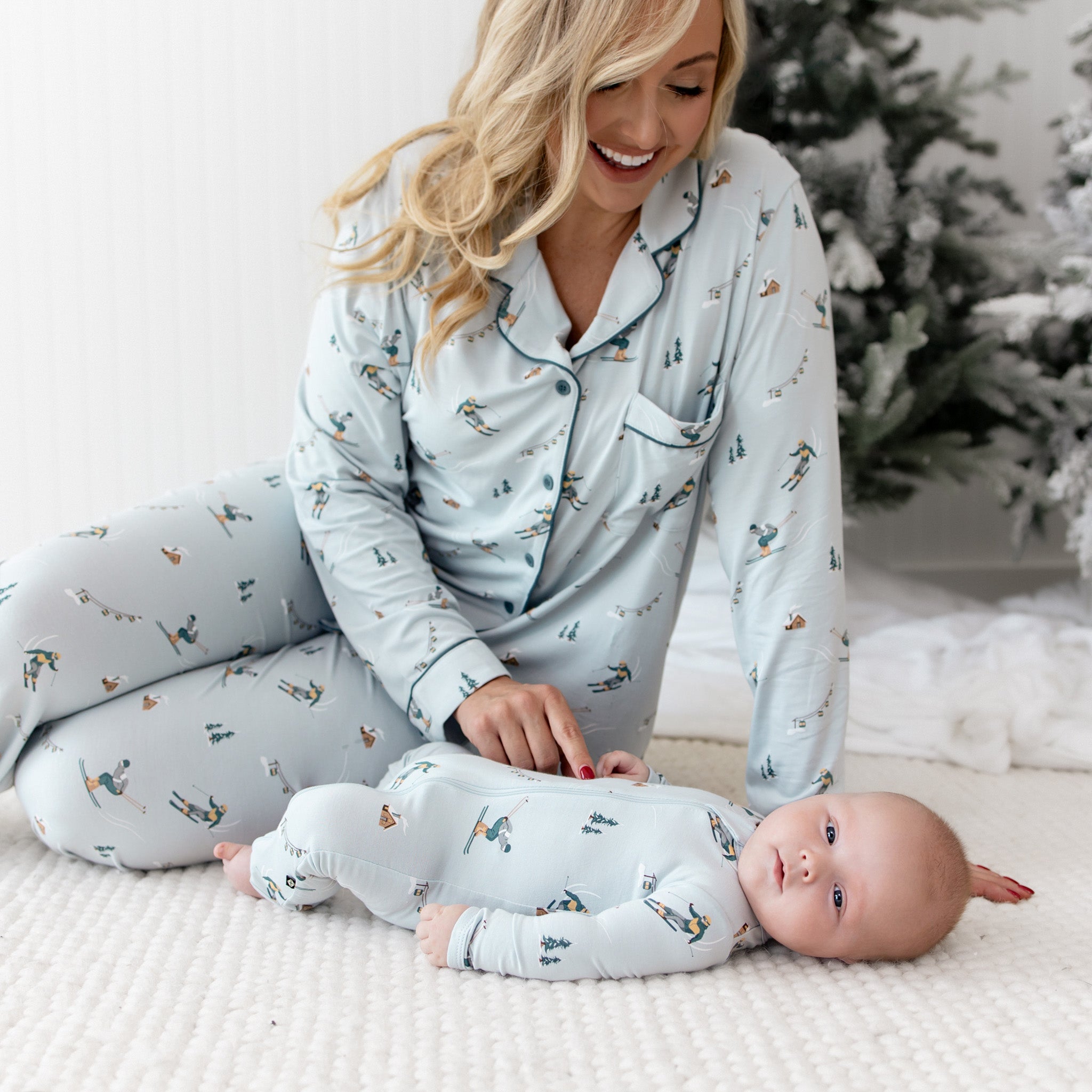 Mother sitting beside her infant who is laying on a white carpet both matching in Ski pajamas