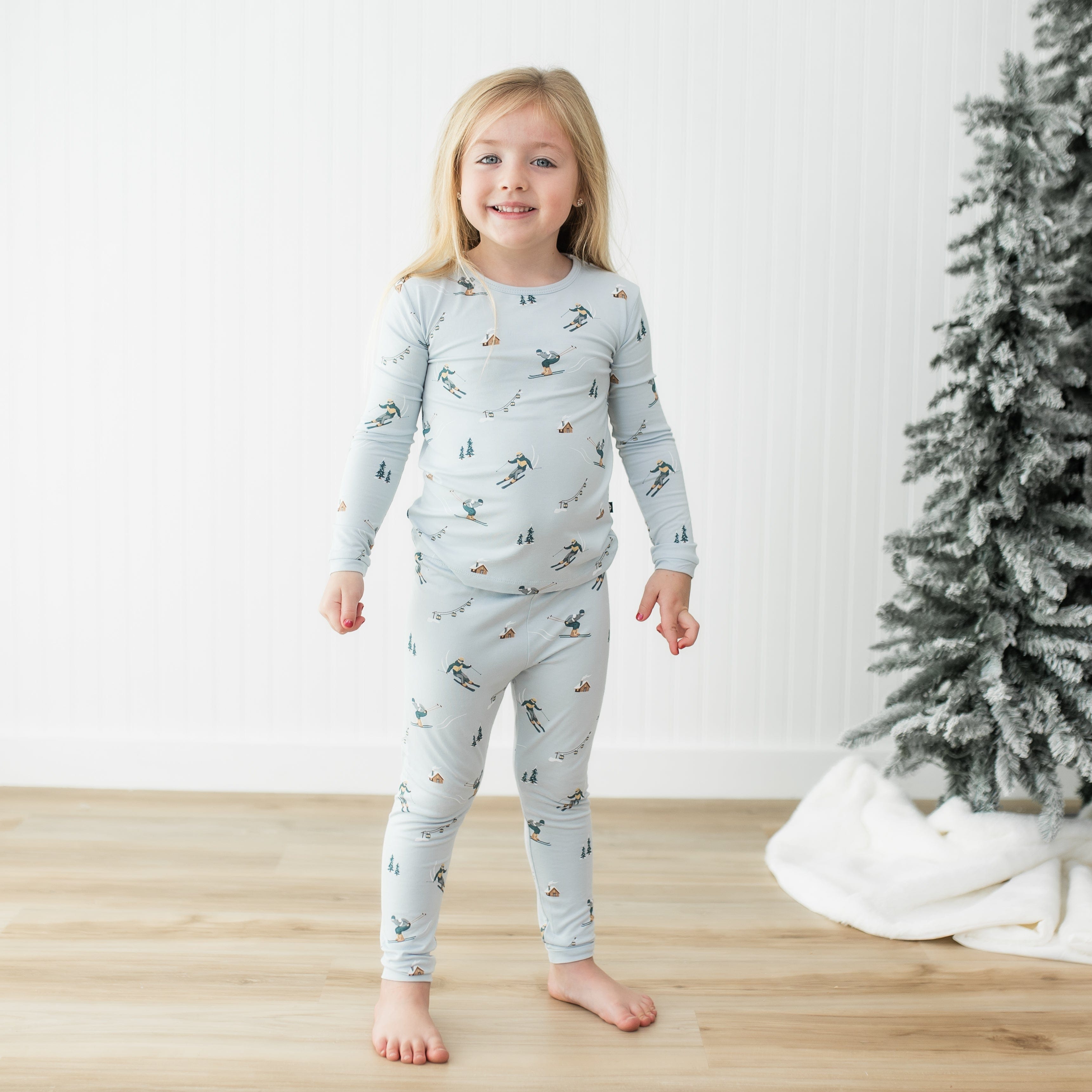 Young girl standing in front of a white paneled wall beside a frosted tree wearing the Long Sleeve Pajamas in Ski