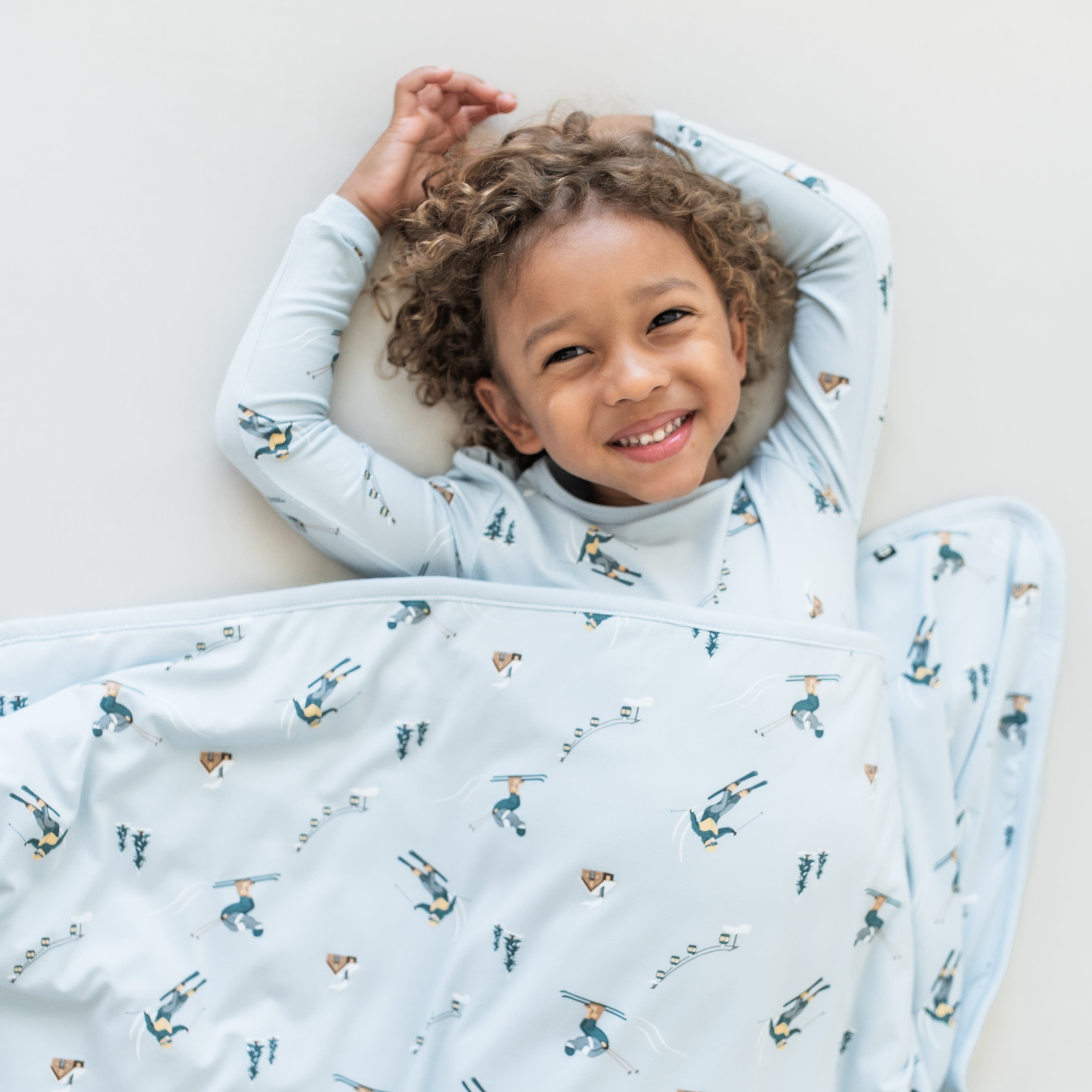 Young smiling child laying on a light colored surface wearing the Long Sleeve Pajamas in Ski with a matching blanket overtop of them