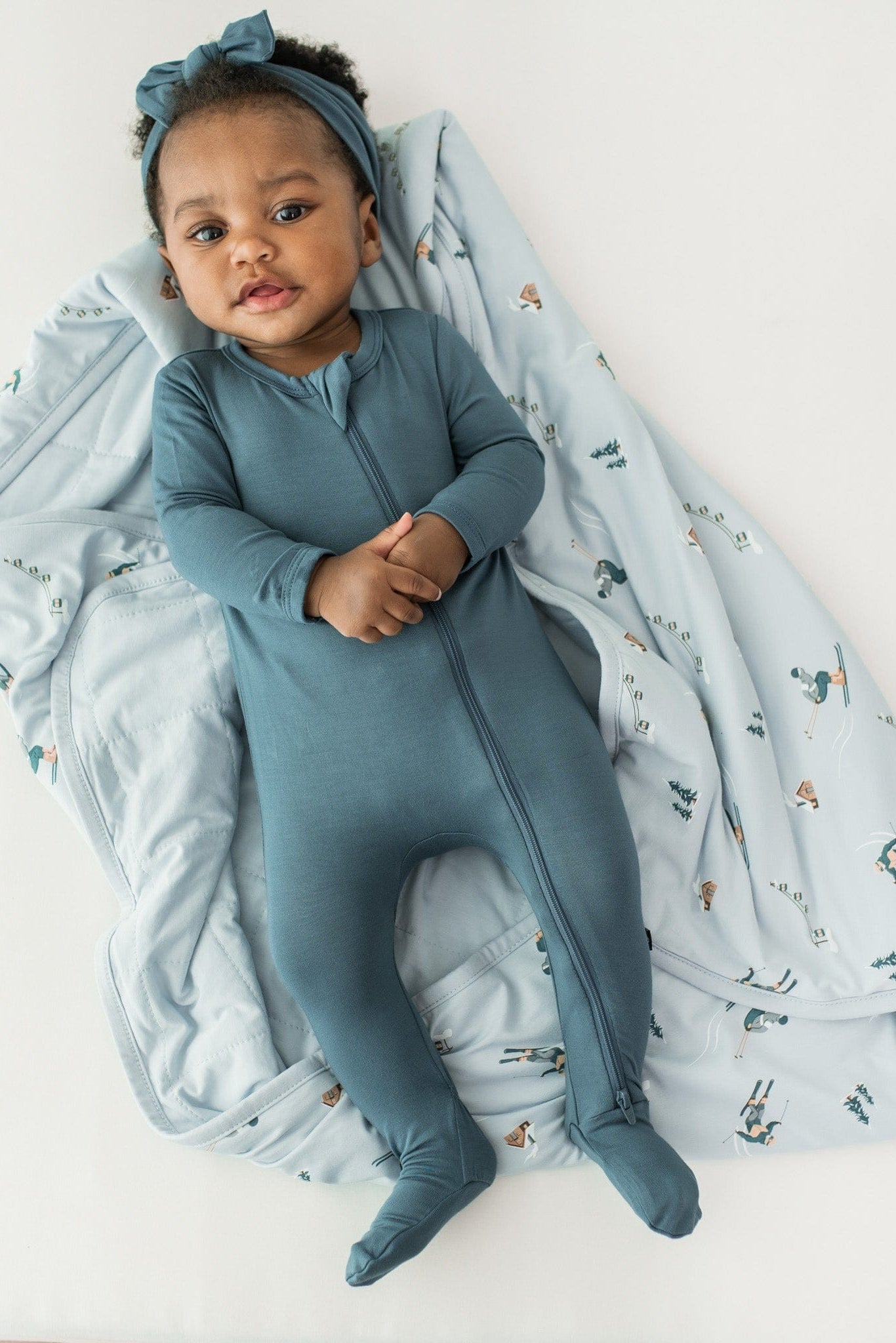 Infant girl laying on the Baby Blanket in Ski wearing a Zippered Footie in Atlantic with a matching bow headband