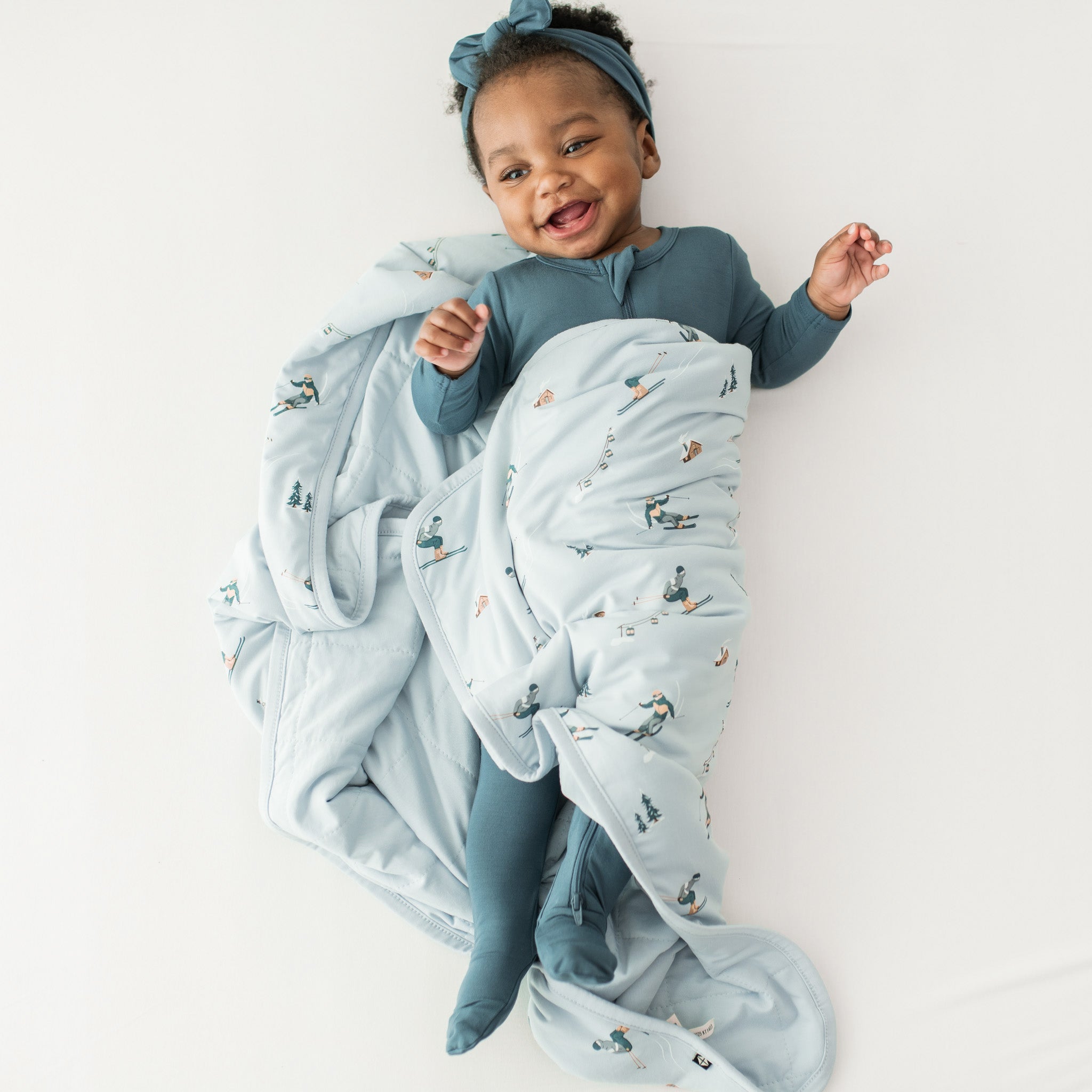 Smiling infant laying down wrapped in a Baby Blanket in Ski wearing a Zippered romper in Atlantic and matching knotted bow headband
