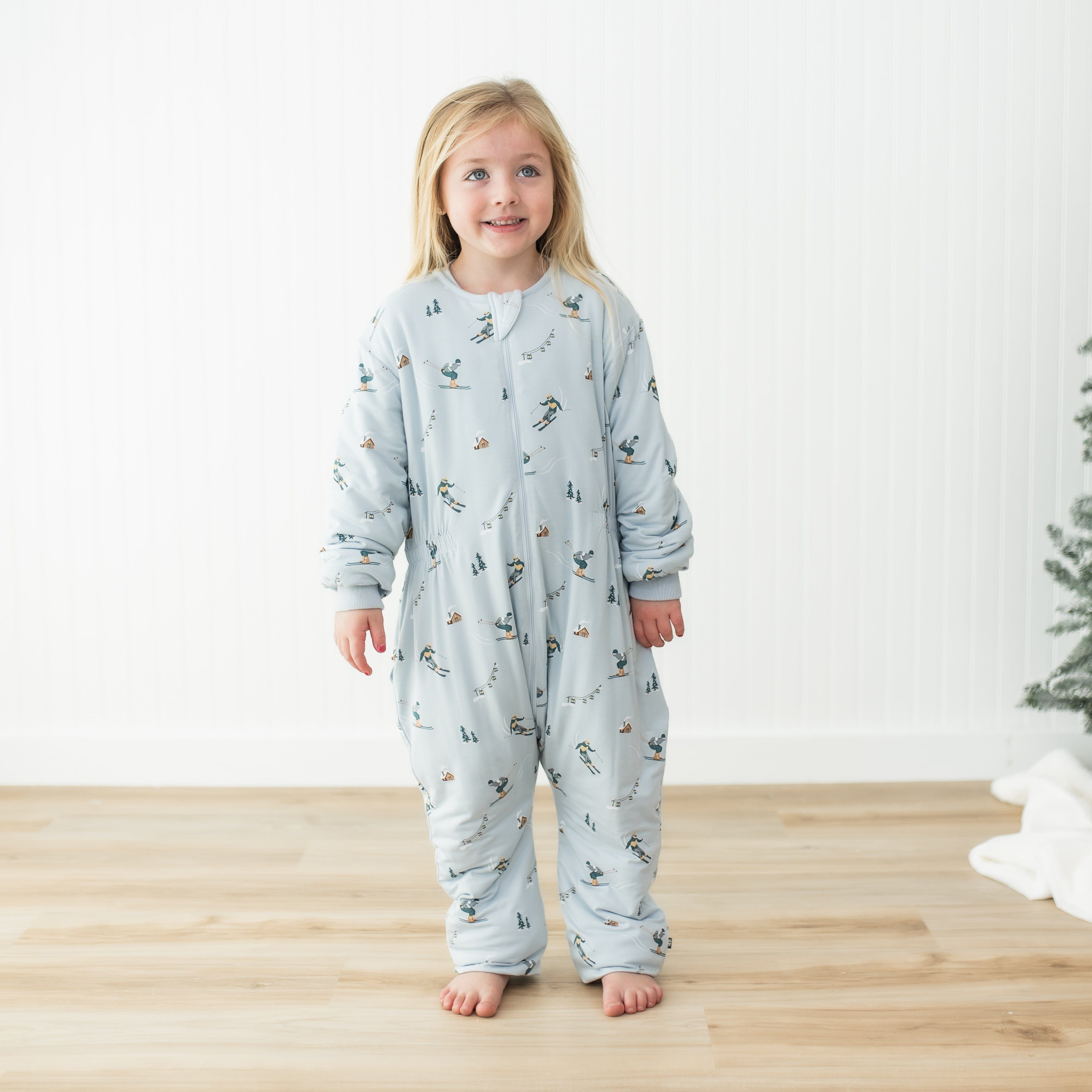 Young girl standing wearing the Cozy Playsuit in Ski on light wood grain floors in front of a white paneled wall