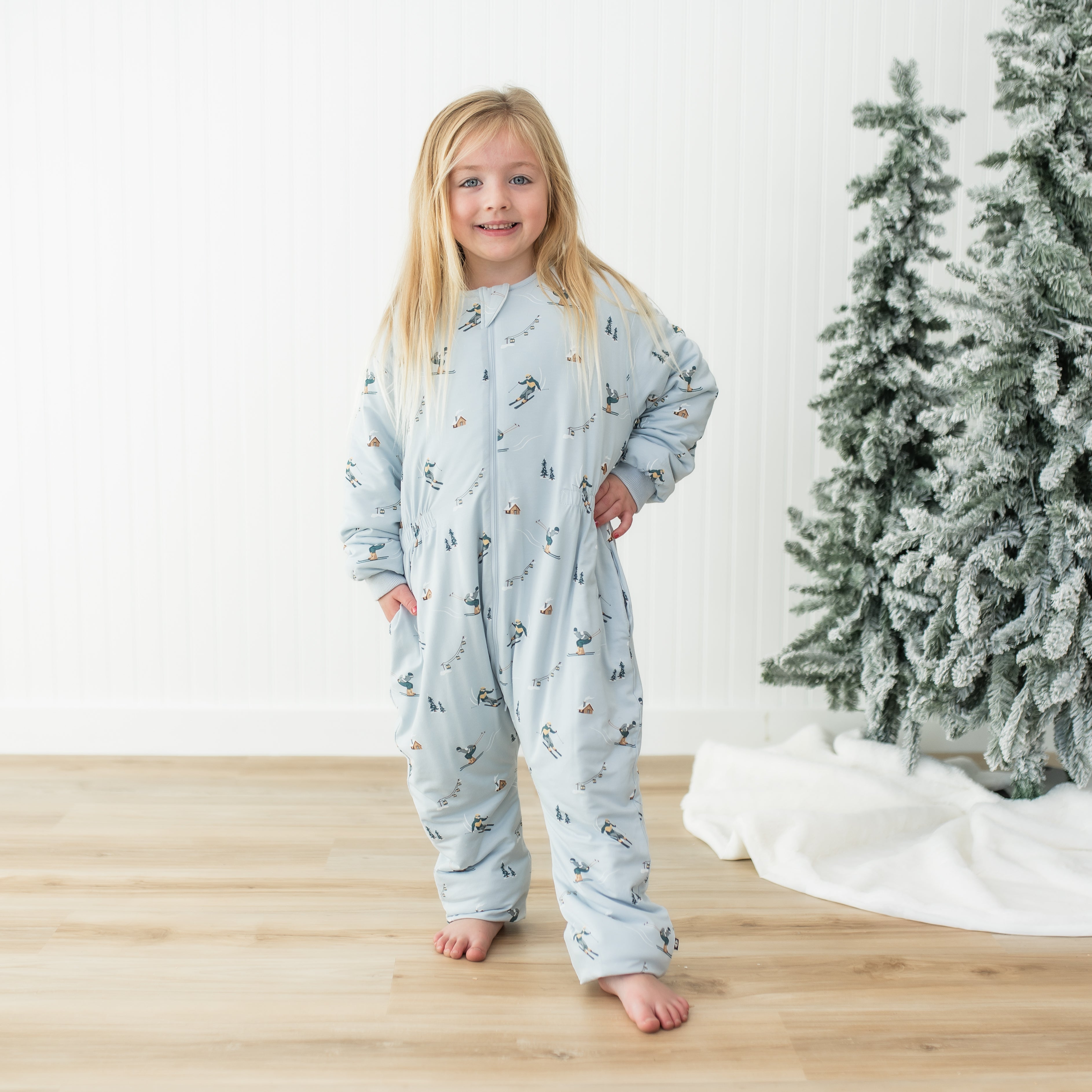 Young girl standing with one hand on her hip wearing the Cozy Playsuit in Ski in front of a white paneled wall and beside a frosted tree