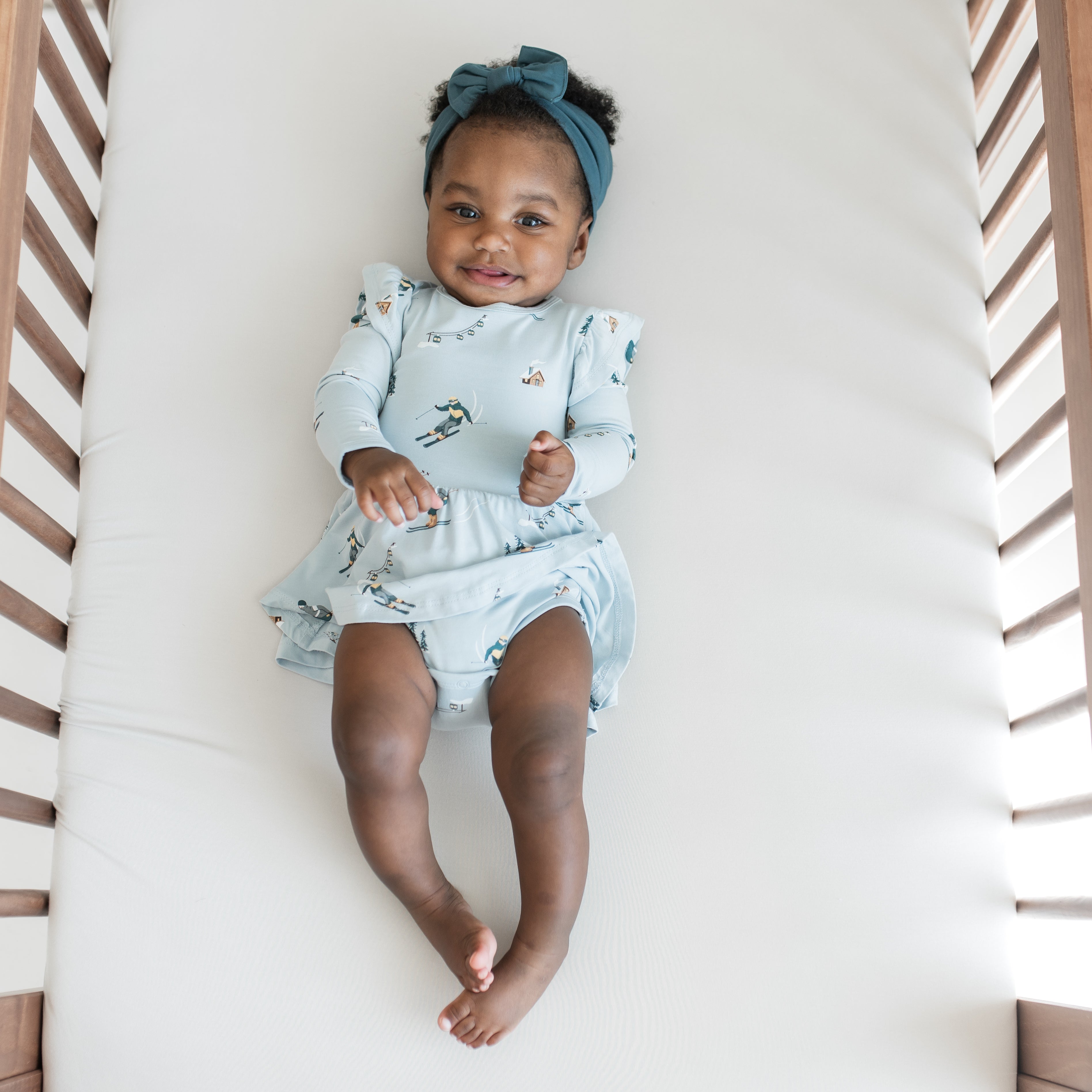Smiling infant laying on a crib wearing the Long Sleeve Twirl Bodysuit Dress in Ski with Atlantic knotted bow headband