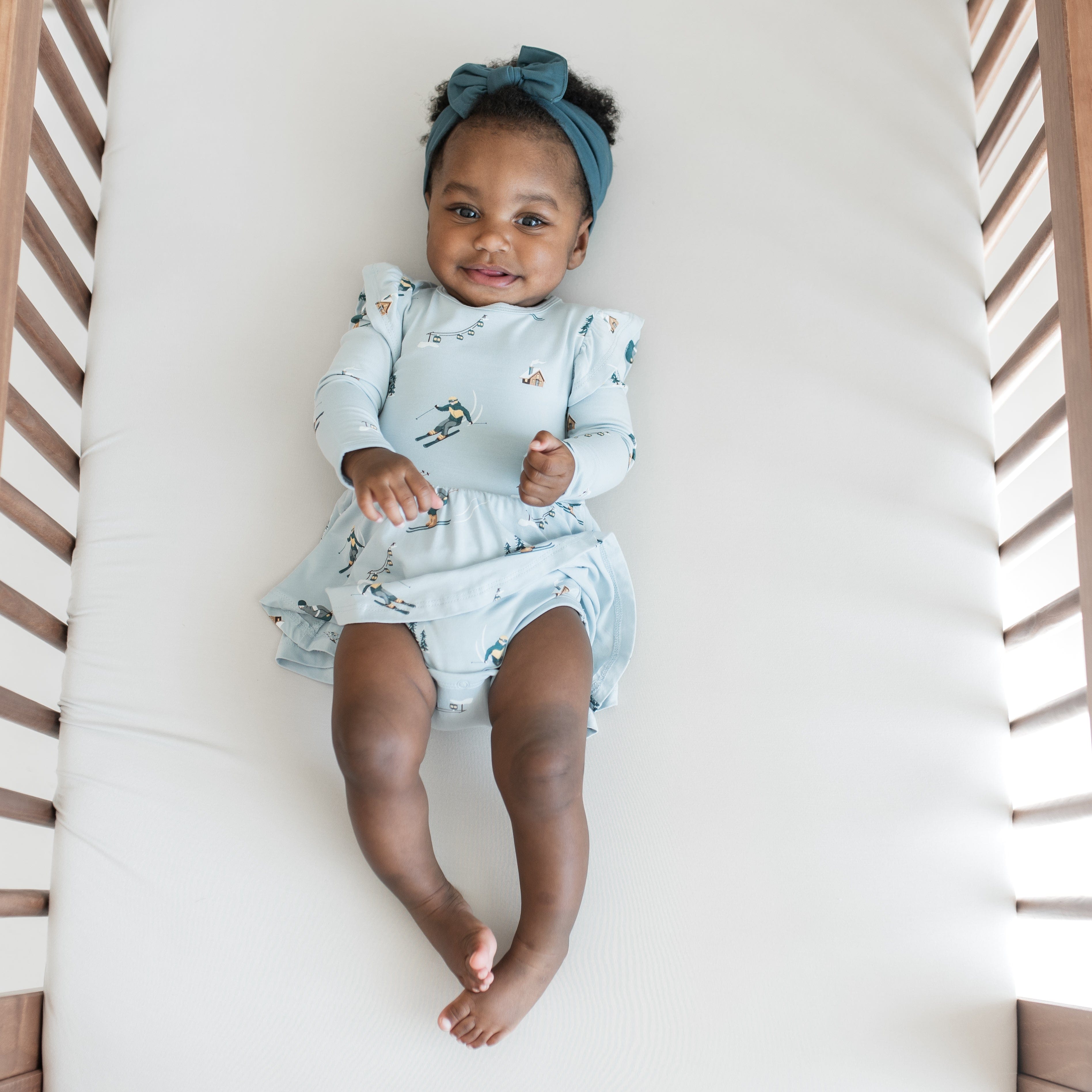 Smiling infant laying on a crib wearing the Long Sleeve Twirl Bodysuit Dress in Ski with Atlantic knotted bow headband