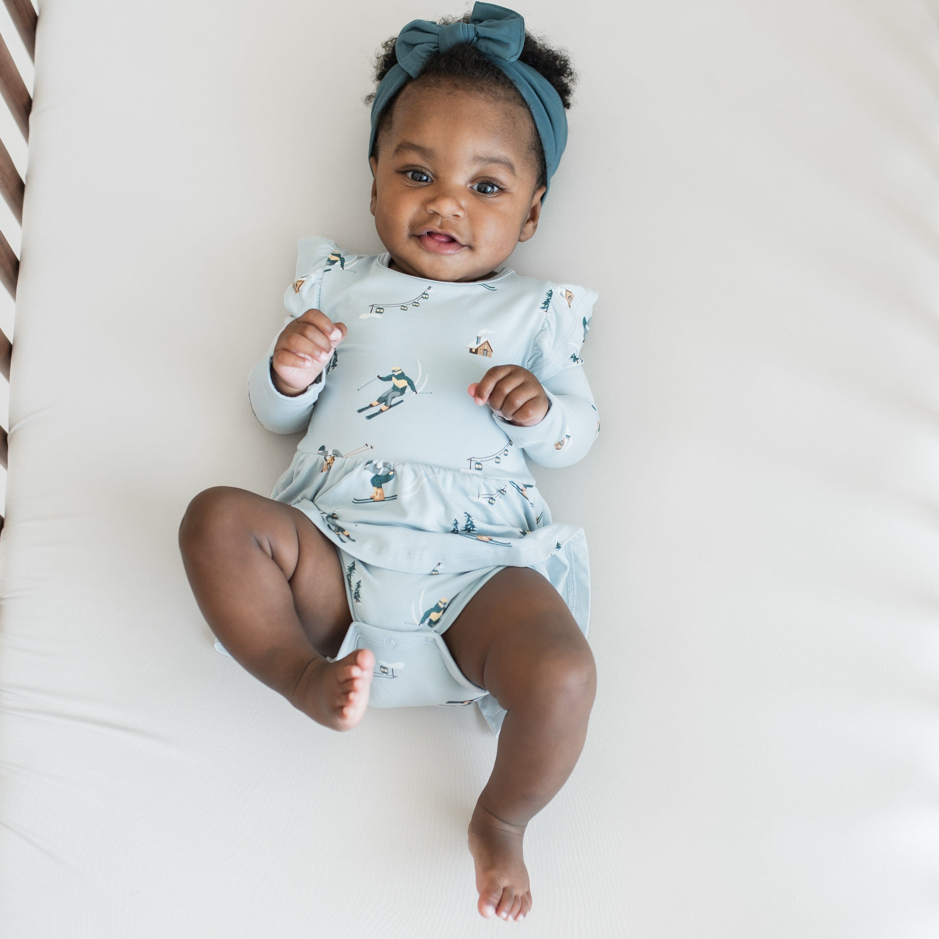 Smiling infant laying in a crib on a light neutral crib sheet wearing the Long Sleeve Twirl Bodysuit Dress in Ski