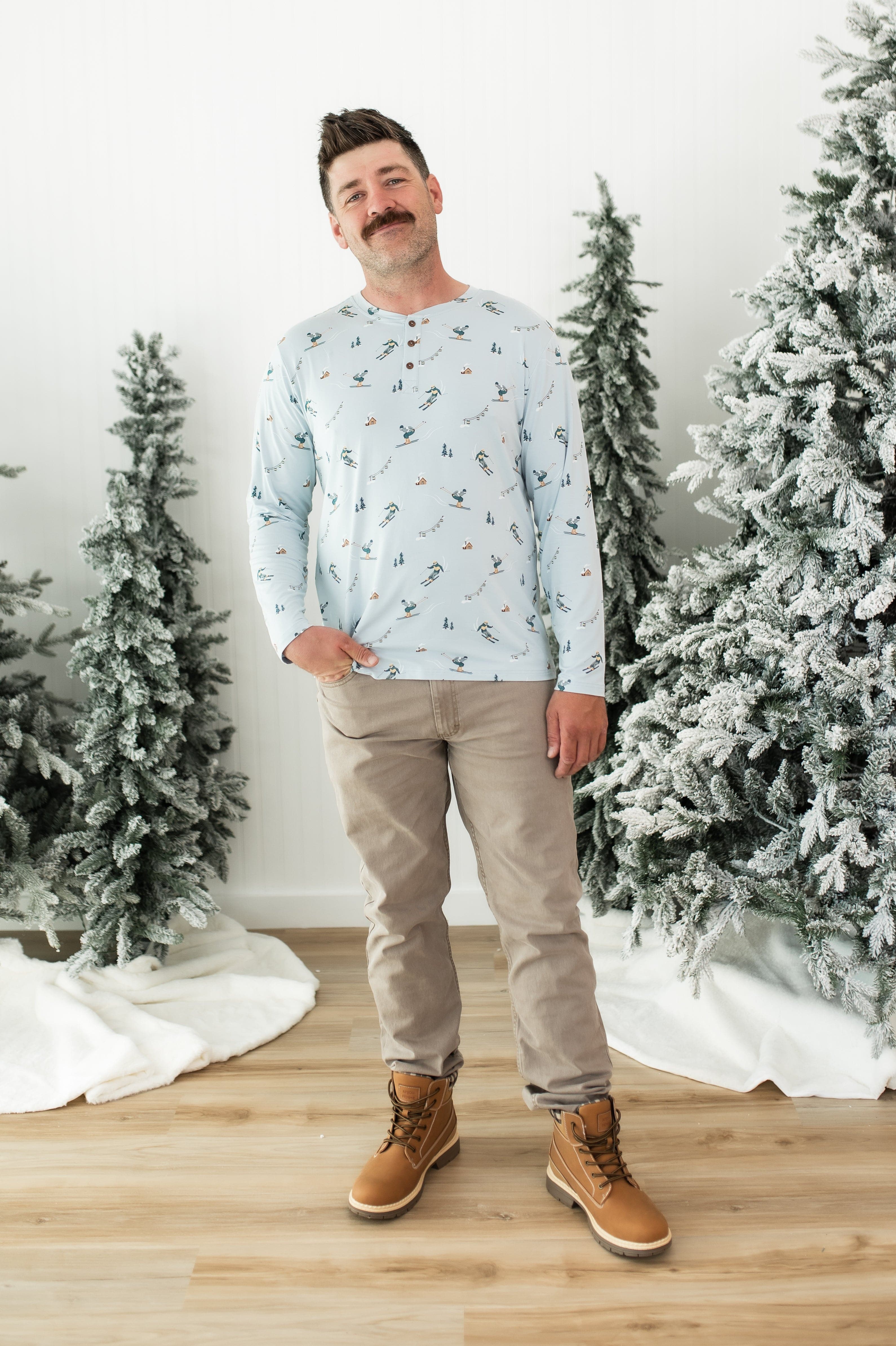 Smiling male modeling the Men's Long Sleeve Henley Top in Ski with khaki colored pants and light brown boots standing between frosted trees