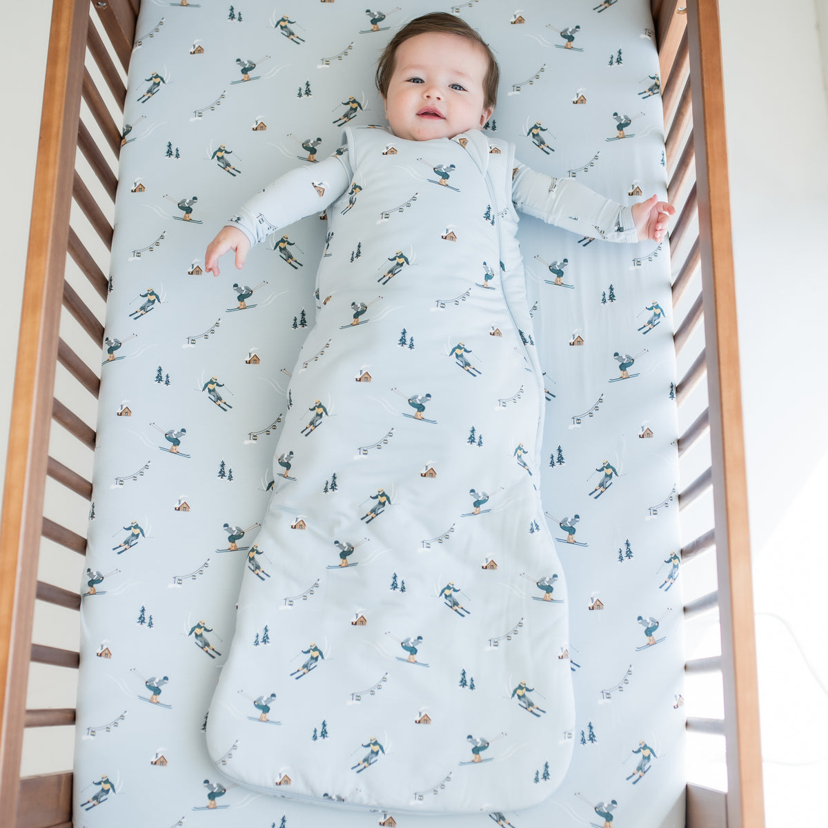Infant laying down in a crib on top of a Ski crib sheet wearing the Sleep Bag in Ski 2.5 with a matching long sleeve romper on underneath