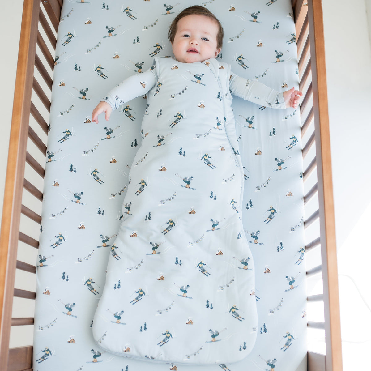 Infant laying down in a crib on top of a Ski crib sheet wearing the Sleep Bag in Ski 2.5 with a matching long sleeve romper on underneath