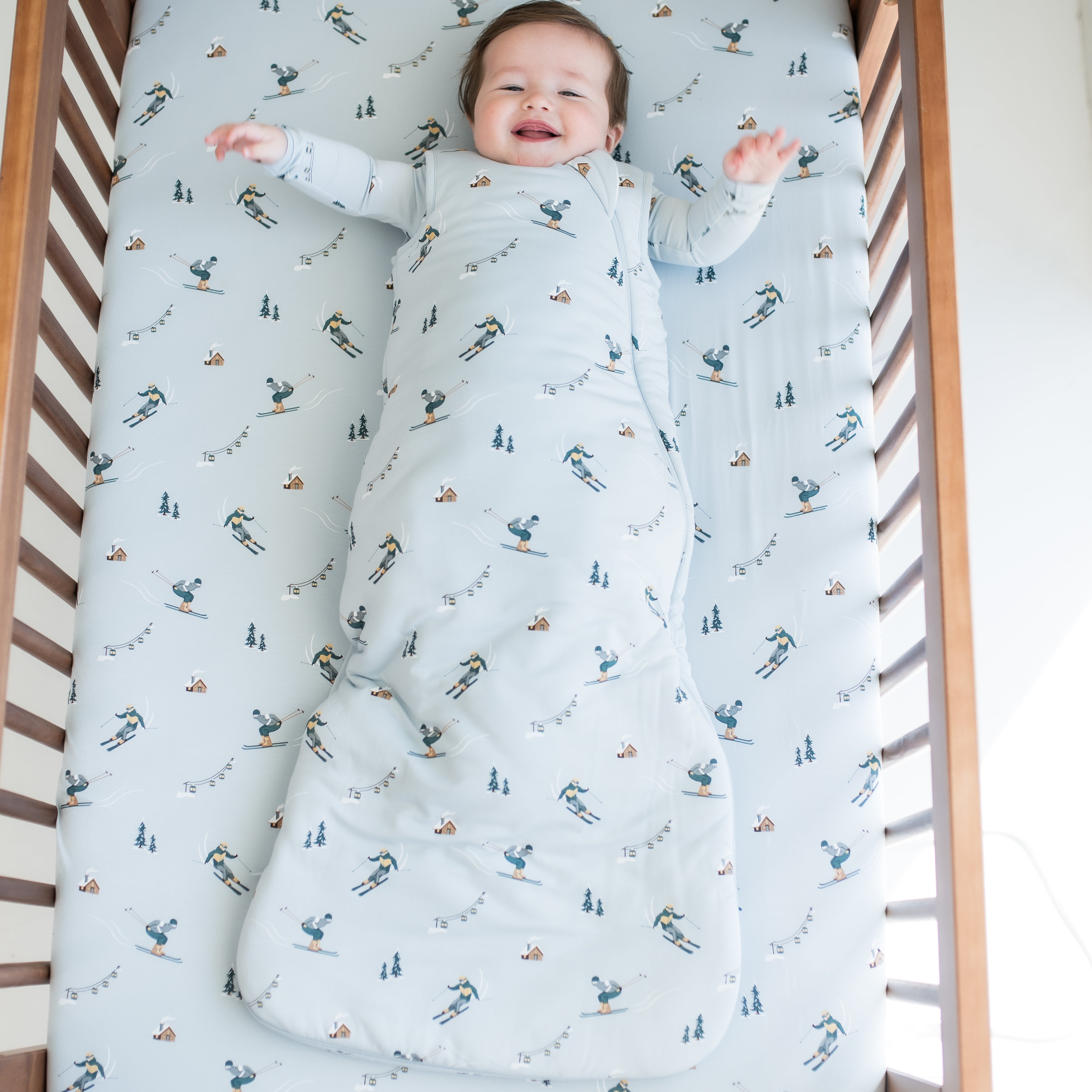 Smiling infant laying in a crib on a ski crib sheet wearing the Sleep Bag in Ski 2.5 with a matching long sleeve romper on underneath