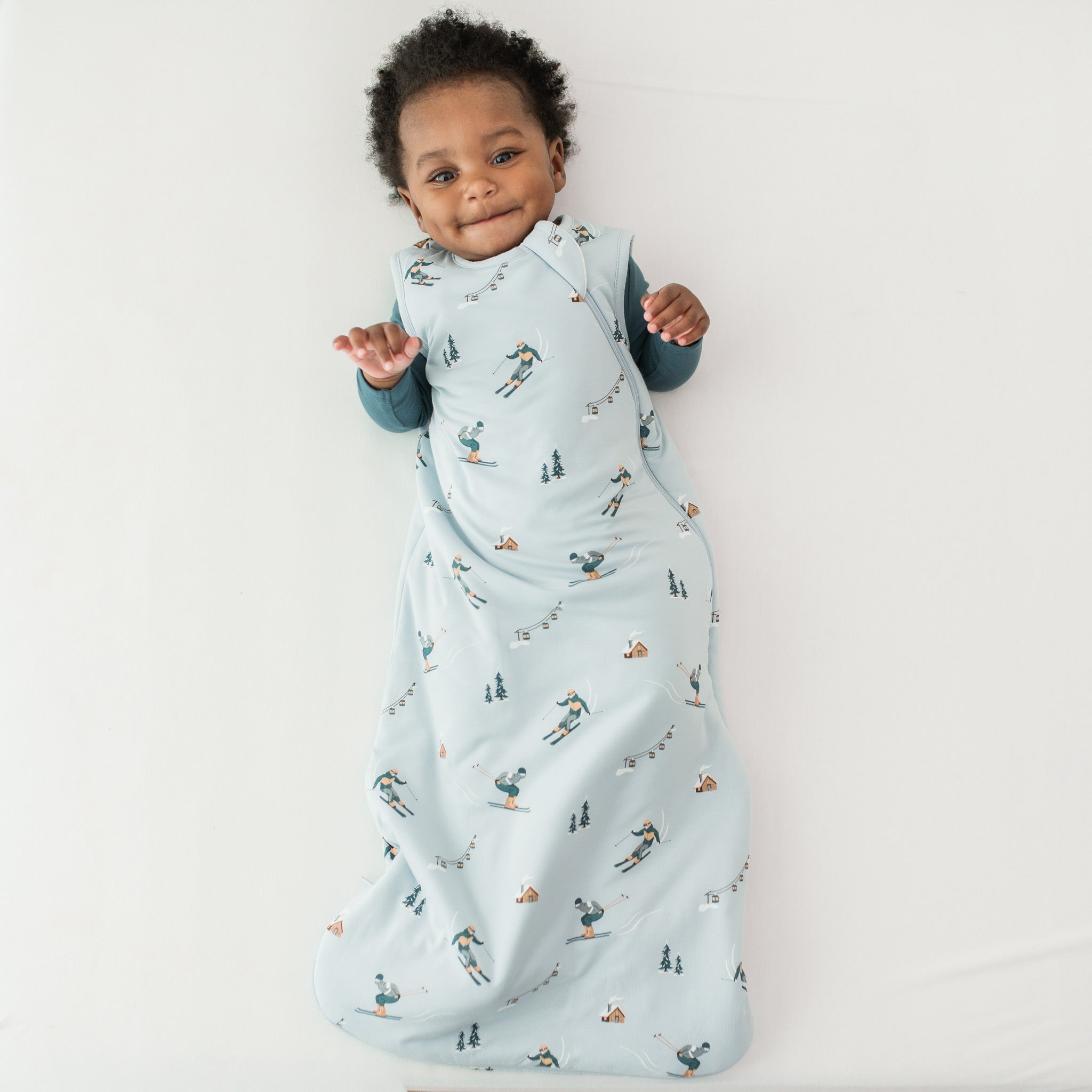 Young toddler laying on a light neutral surface wearing the Sleep Bag in Ski 1.0 with an Atlantic Romper underneath