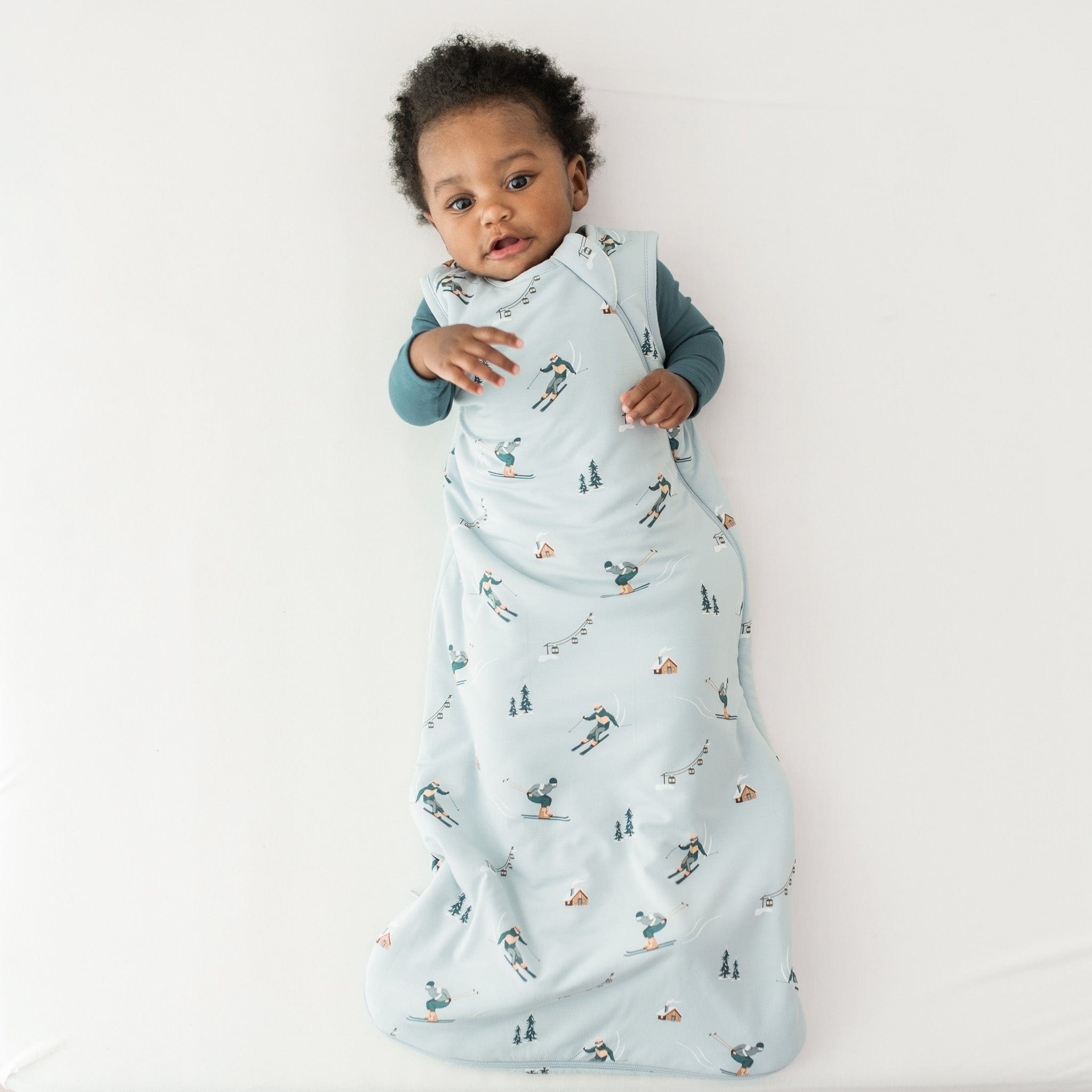 Young toddler laying on a light neutral surface wearing the Sleep Bag in Ski 1.0 with an Atlantic romper on underneath