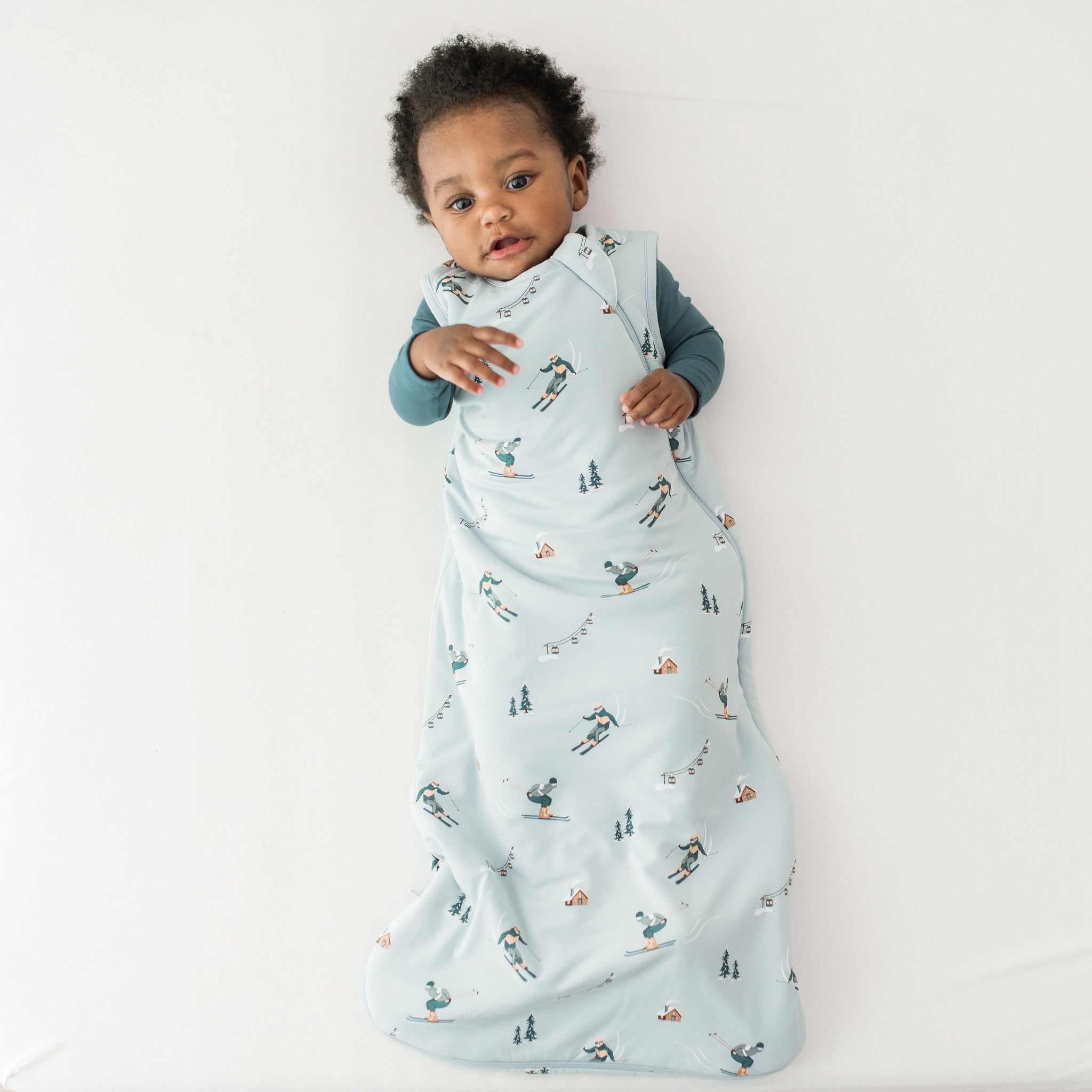 Young toddler laying on a light neutral surface wearing the Sleep Bag in Ski 1.0 with an Atlantic romper on underneath